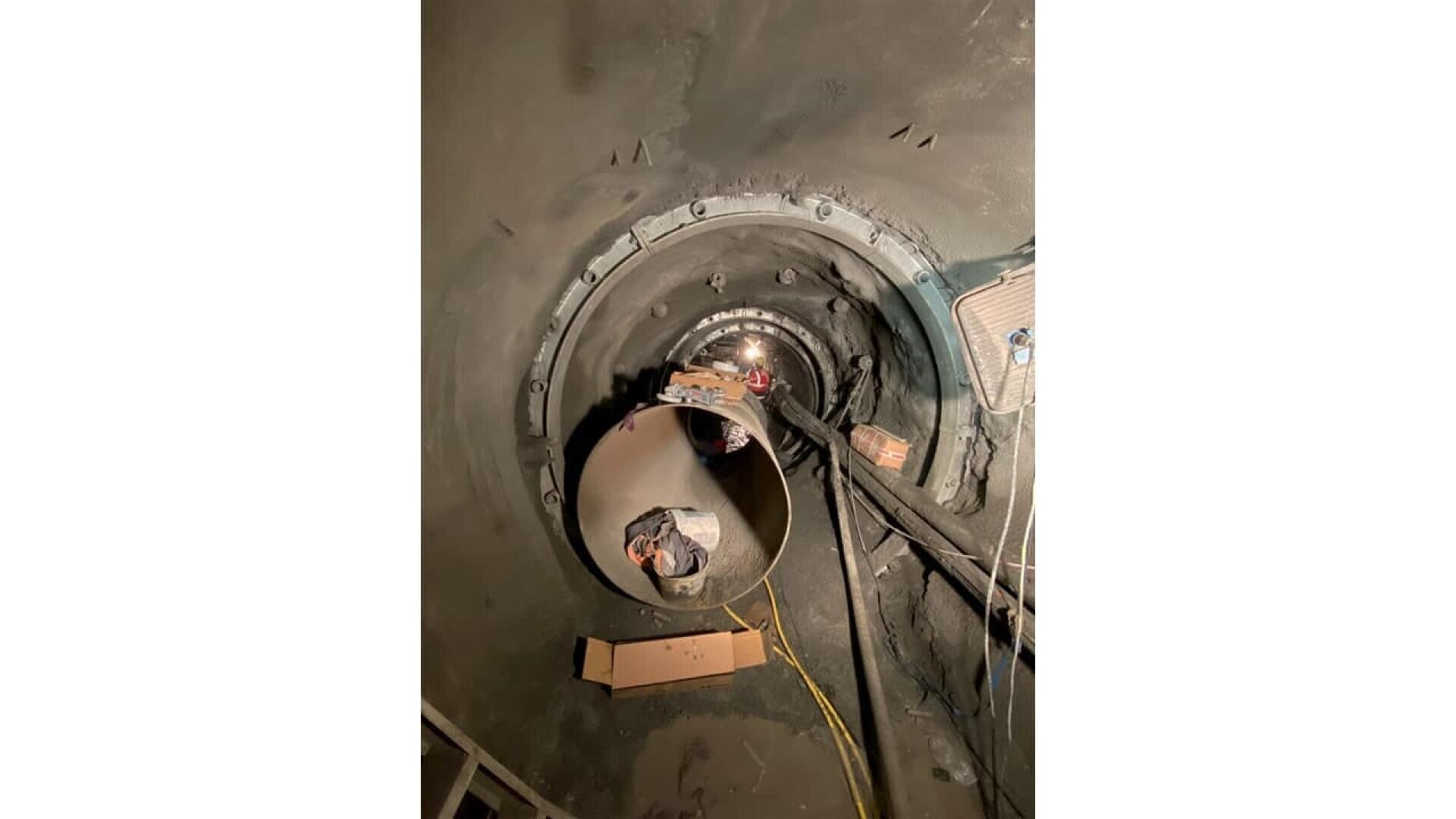 A close-up view inside a large industrial pipe or chamber, showing cables, wires, and a reflective spherical component at the center, with tools and a cardboard box nearby. The surroundings appear metallic and industrial.
