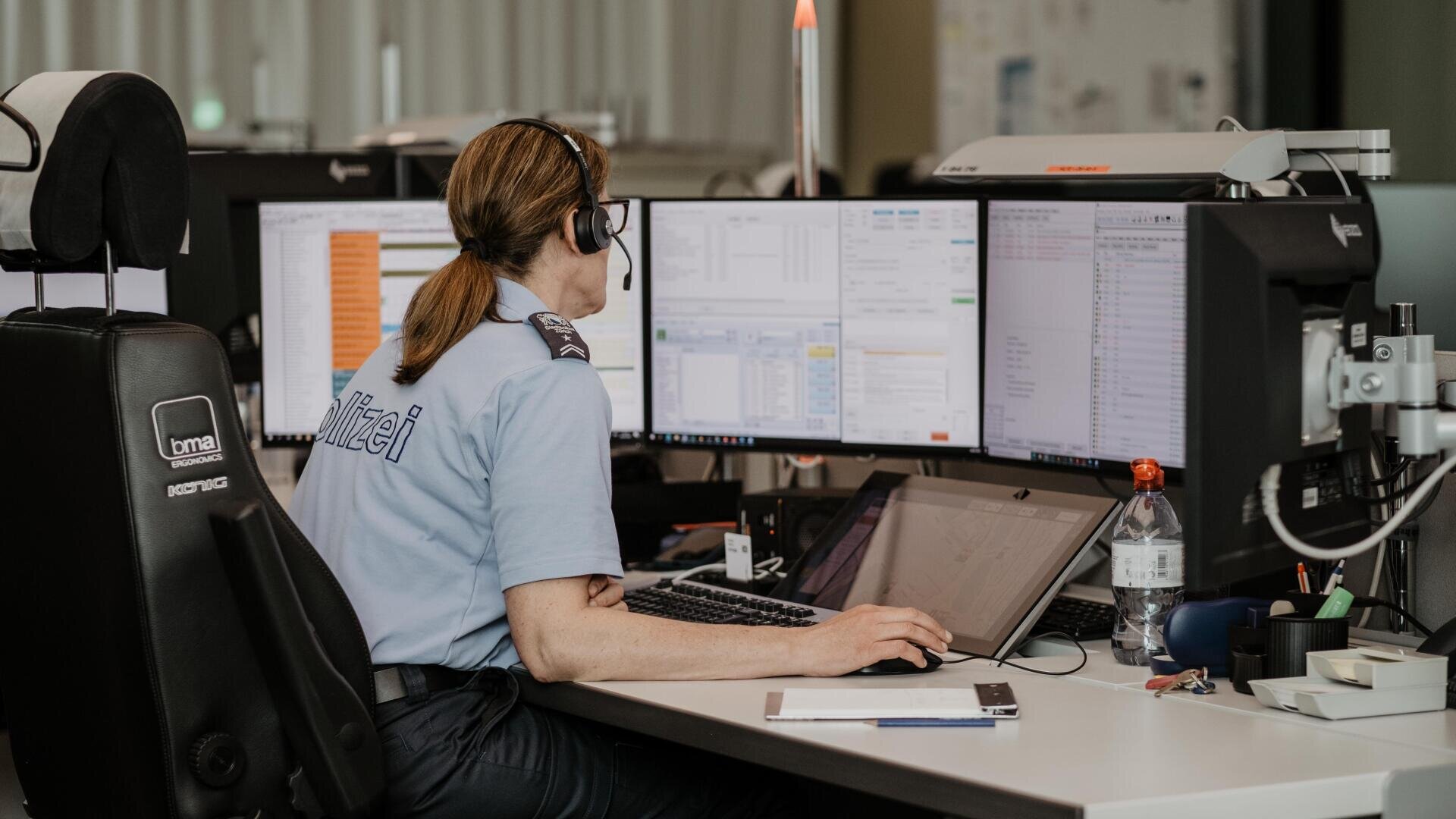 A police dispatcher wearing a headset sits at a desk with multiple monitors displaying data and screens, working in a modern office environment. A water bottle and office supplies are nearby.