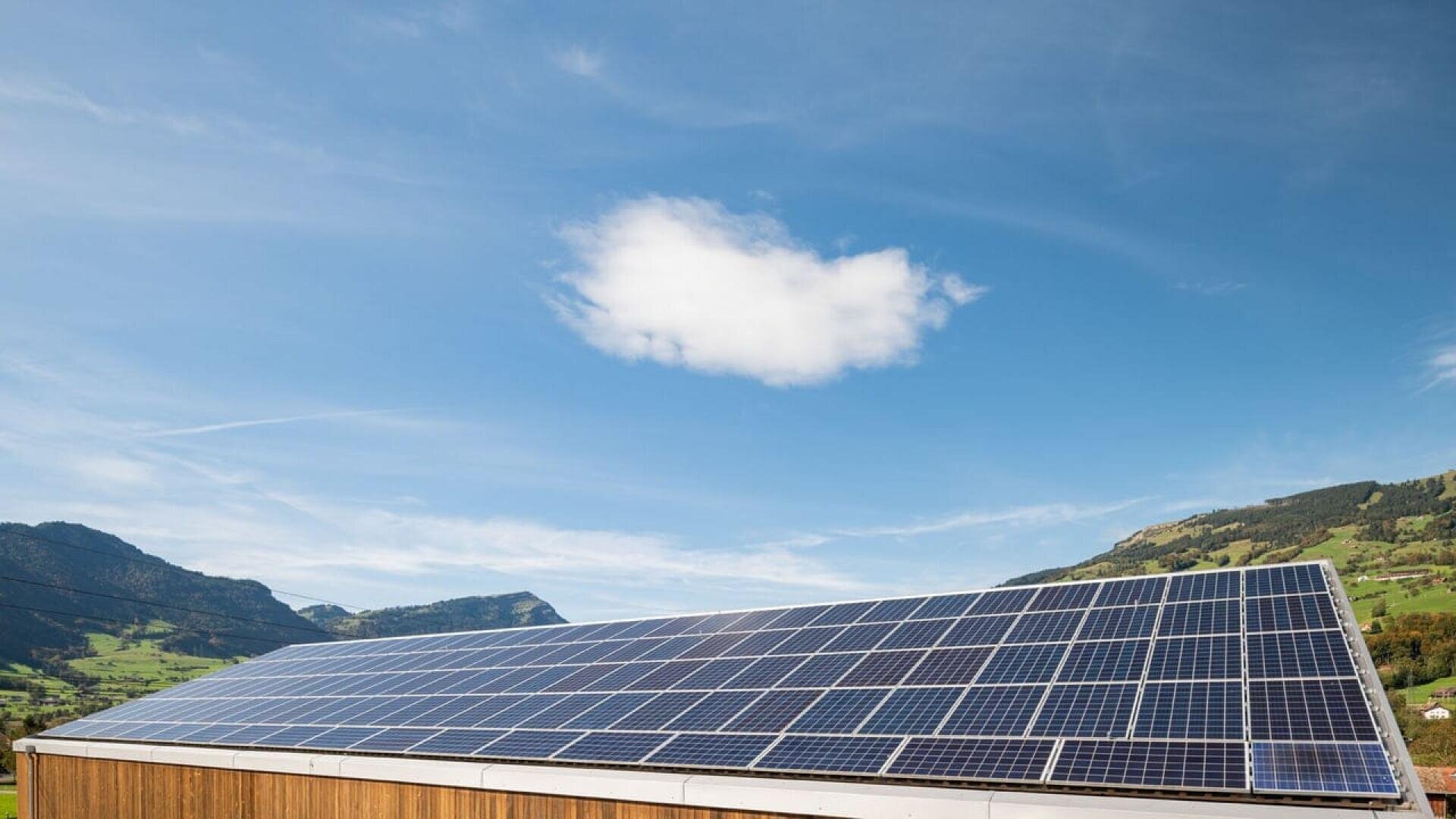 A rooftop covered with solar panels under a clear blue sky with a single white cloud, surrounded by green hills and distant mountains.