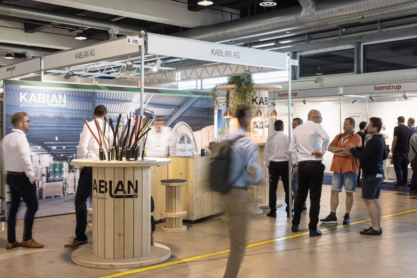 In einer hell erleuchteten Messehalle umrunden die Menschen einen Messestand der KABLAN AG, der mit Industriekabeln auf einer Holzspule, Bannern und Holzdekor ausgestattet ist.