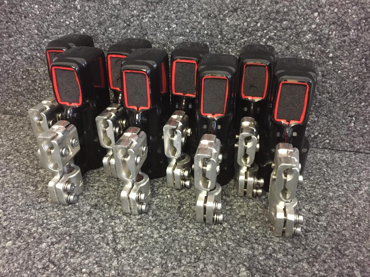 Sechs schwarze mechanische Geräte mit rot umrandeten quadratischen Abschnitten und angebrachten silbernen Metallteilen, die in zwei Reihen auf einer strukturierten grauen Oberfläche angeordnet sind.