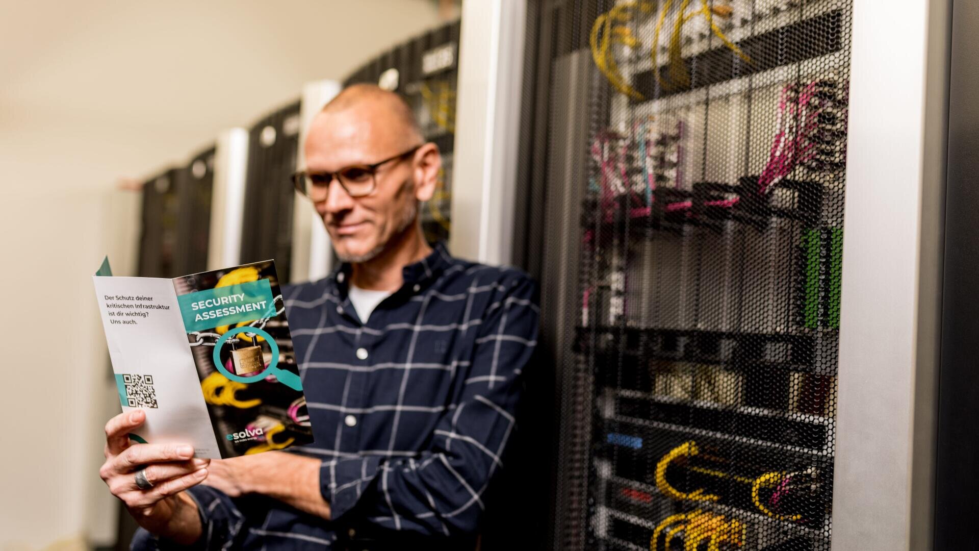 Eine Person mit Brille und kariertem Hemd steht in einem Serverraum und liest eine Broschüre zur Sicherheitsbewertung. Die Serverschränke hinter ihnen sind mit Kabeln und Geräten gefüllt.