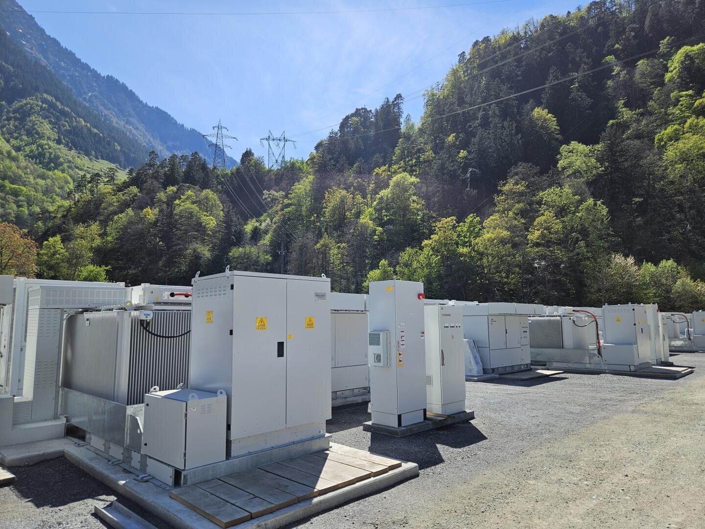 Rows of white electrical appliances and storage units stand outside on a gravel surface, surrounded by green, wooded mountains under a clear blue sky with power lines visible above.