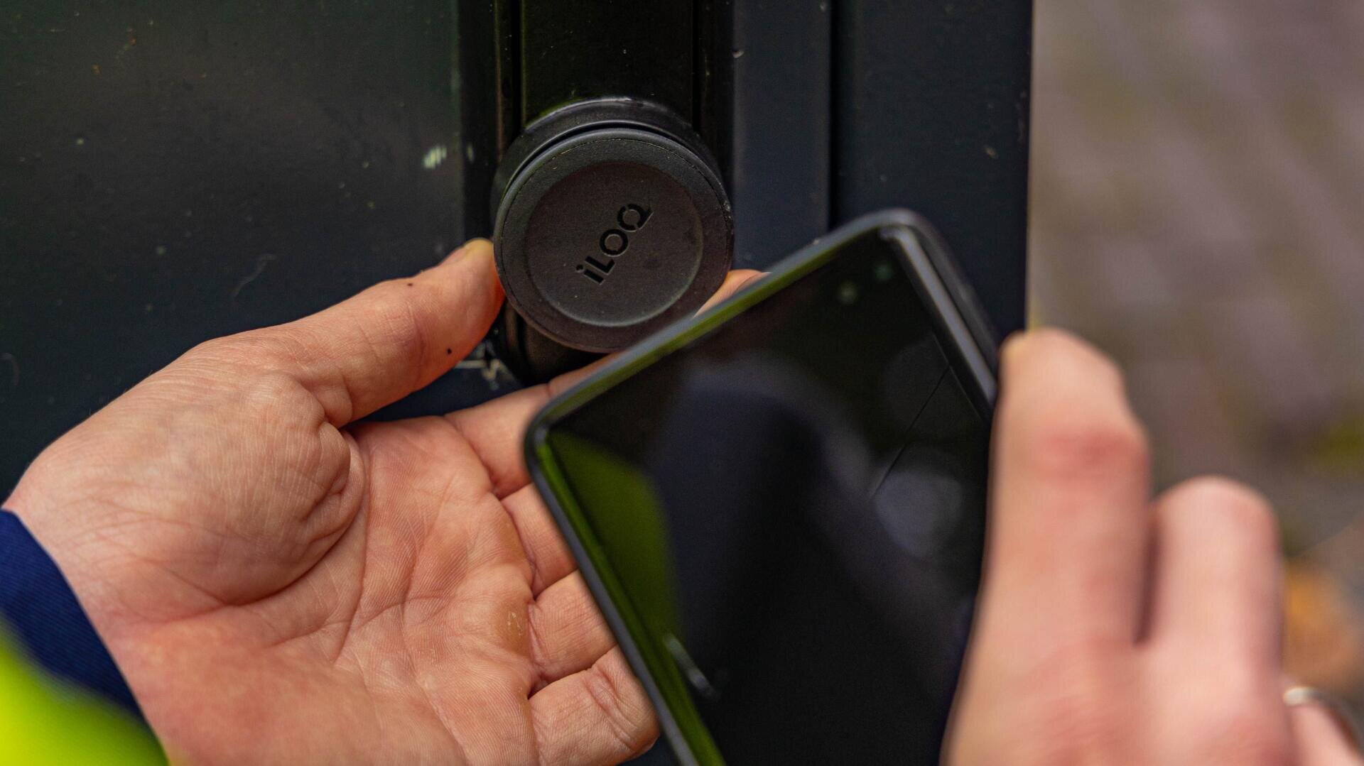A person holds a phone close to a round black iLOQ smart lock on a door, appearing to use the phone to unlock it.