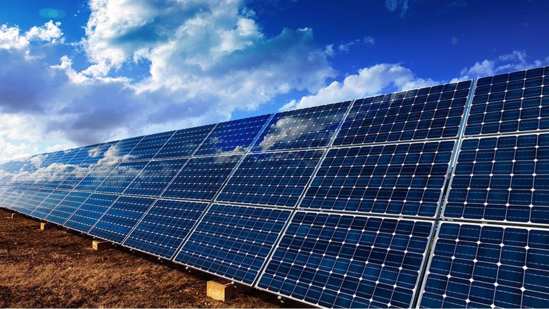 Rows of solar panels installed outdoors on a grassy field, reflecting the blue sky and clouds above, with a bright sunlight illuminating the scene.