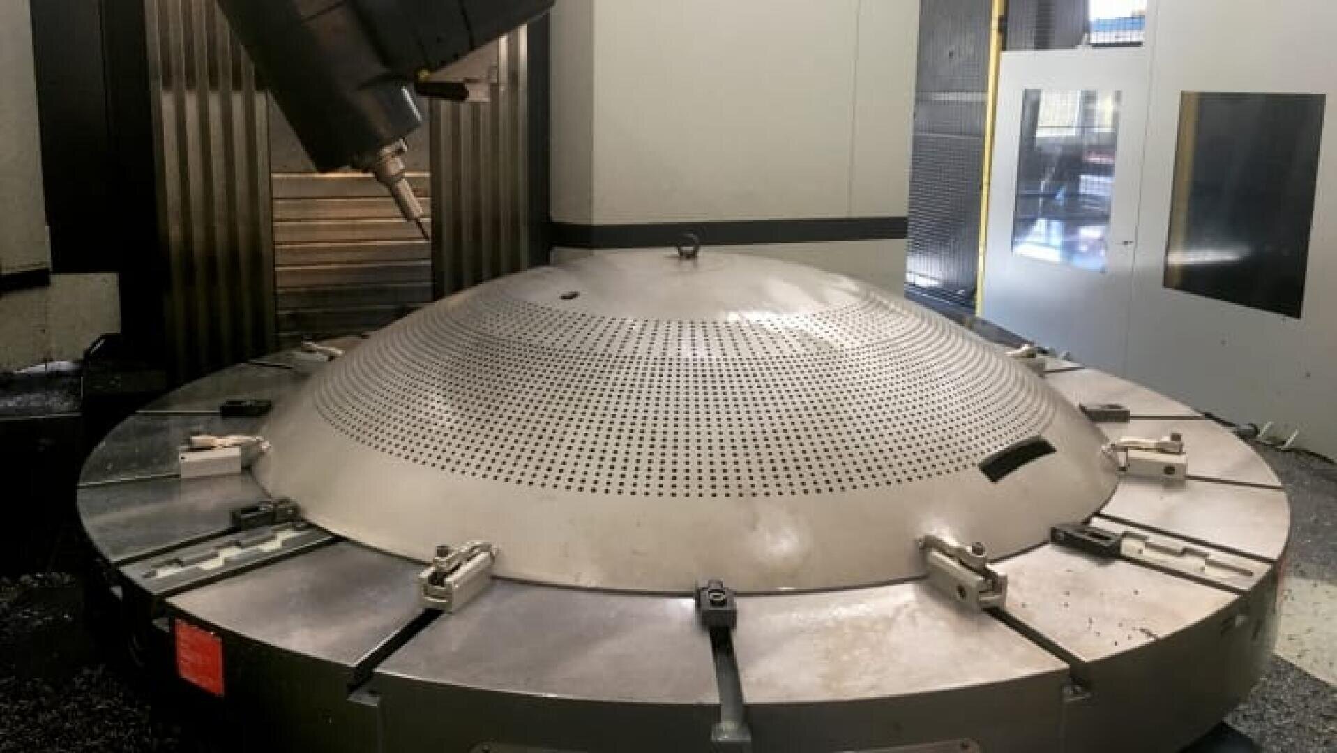 Ein großes, rundes Metallteil mit vielen kleinen Löchern ist auf einem Maschinenbett befestigt. Darüber befindet sich ein industrieller Werkzeugkopf, wahrscheinlich für Präzisionsbohrungen oder -bearbeitungen in einer High-Tech-Fertigungsanlage.
