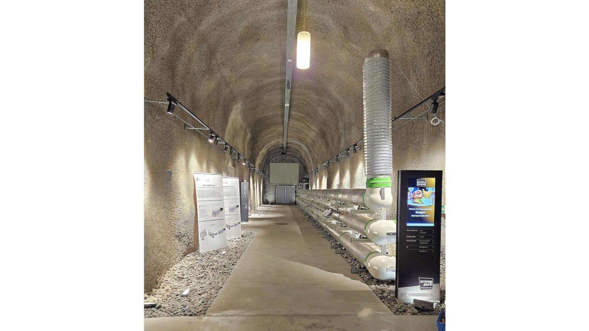 An underground tunnel with arched ceilings contains display boards and a row of large pipes and tanks along the right wall, illuminated by overhead and side lights. Gravel lines the sides of the concrete walkway.