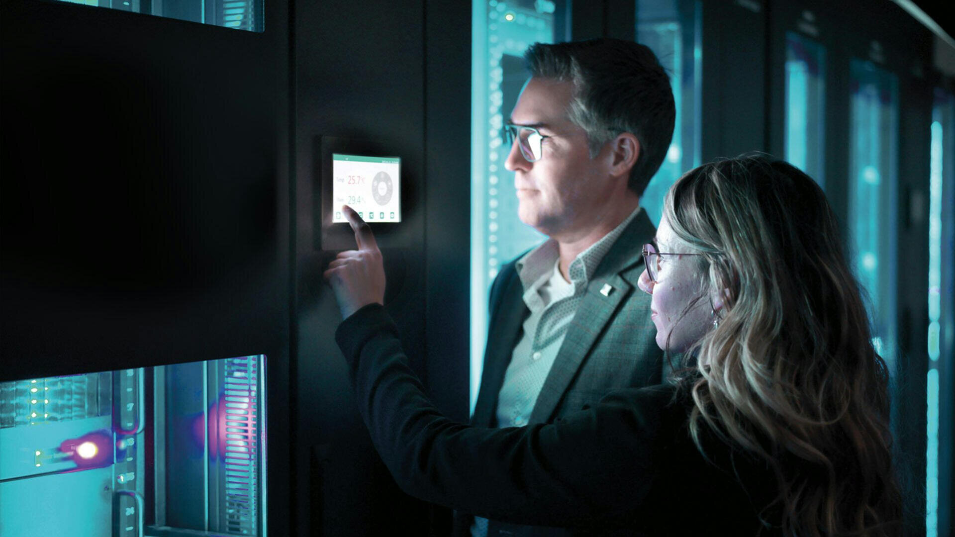 Zwei Personen stehen in einem Serverraum und untersuchen ein Touchscreen-Panel auf einem Serverschrank und interagieren damit. Der Raum wird von der kühlen blauen Beleuchtung der Serverausrüstung beleuchtet.