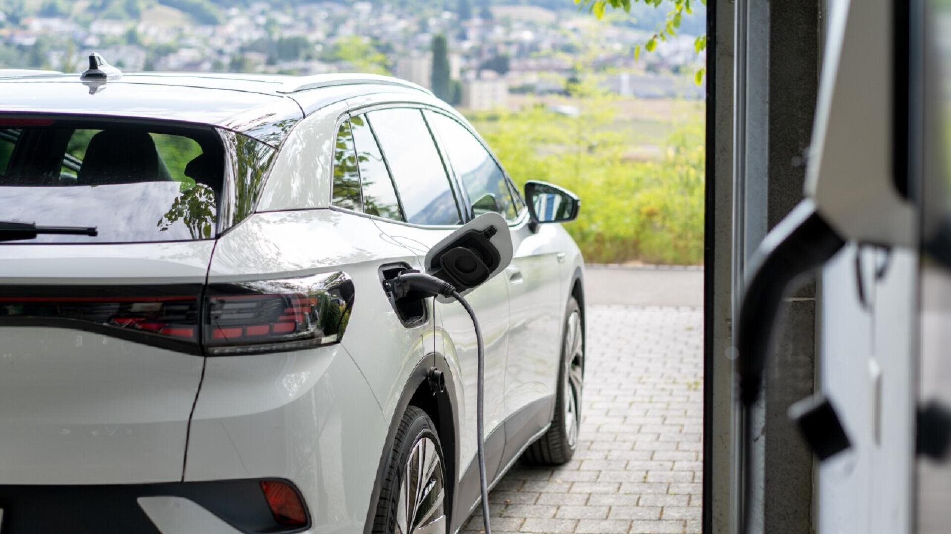 A white electric car is parked outdoors and charging, with the charging cable plugged into its side. The scene is bright and leafy, with a blurred cityscape visible in the background.