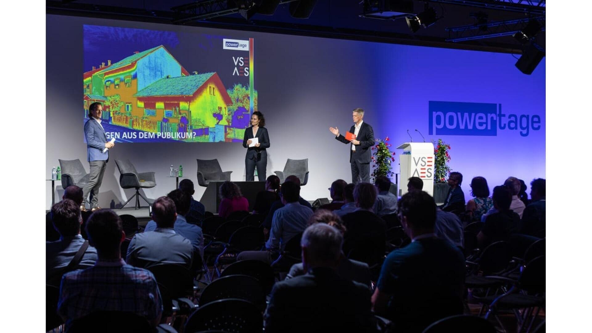 A conference or seminar with three speakers on stage, an audience seated facing them, and a large screen displaying a colorful thermal image of buildings. The word powertage is visible on the wall.