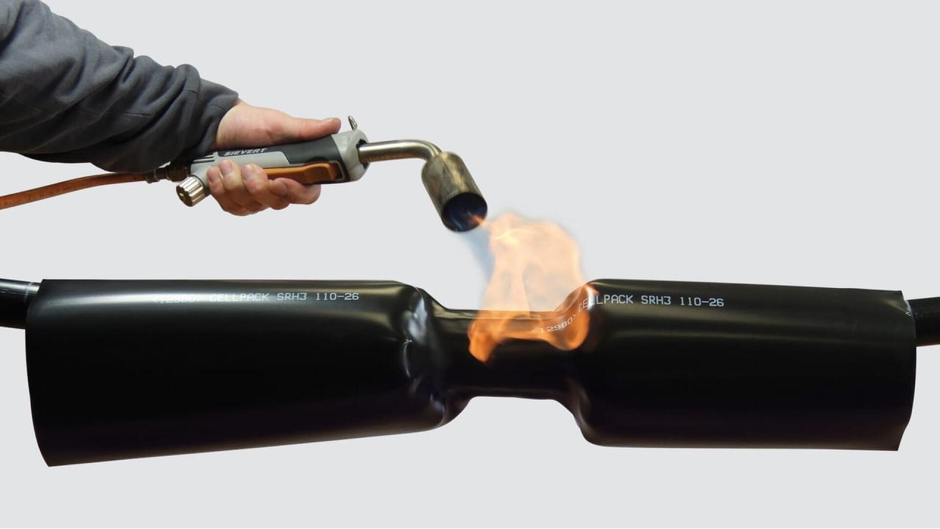 A person using a handheld blowtorch to heat and shrink a black heat-shrink sleeve around a pipe, creating a secure seal.