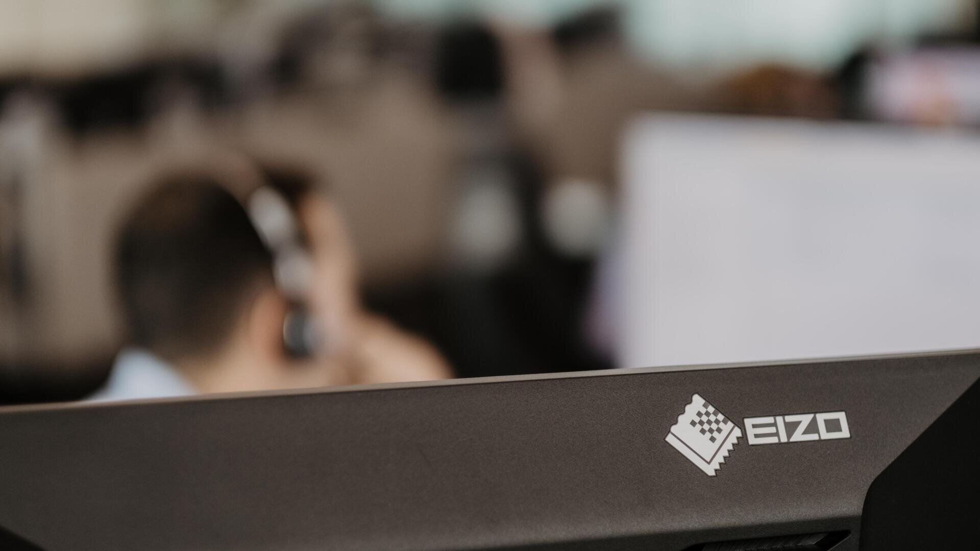 Nahaufnahme eines EIZO-Computermonitors in einer Büroumgebung, mit einem unscharfen Hintergrund, der eine Person mit einem Headset zeigt, was auf eine Arbeits- oder Call-Center-Umgebung hindeutet.