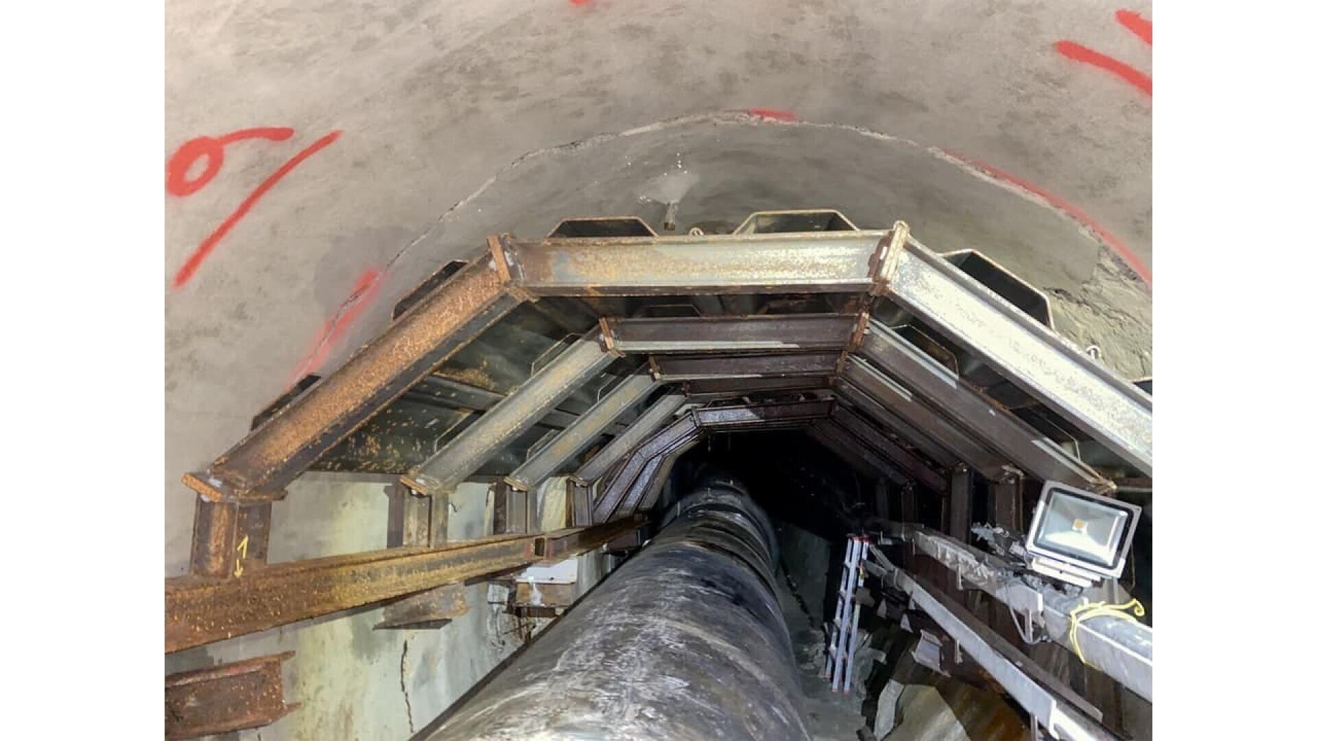 View looking down a narrow, reinforced concrete tunnel lined with rusted metal beams, a large black pipe running through the center, and a mounted work light on the right; red markings on the tunnel wall.