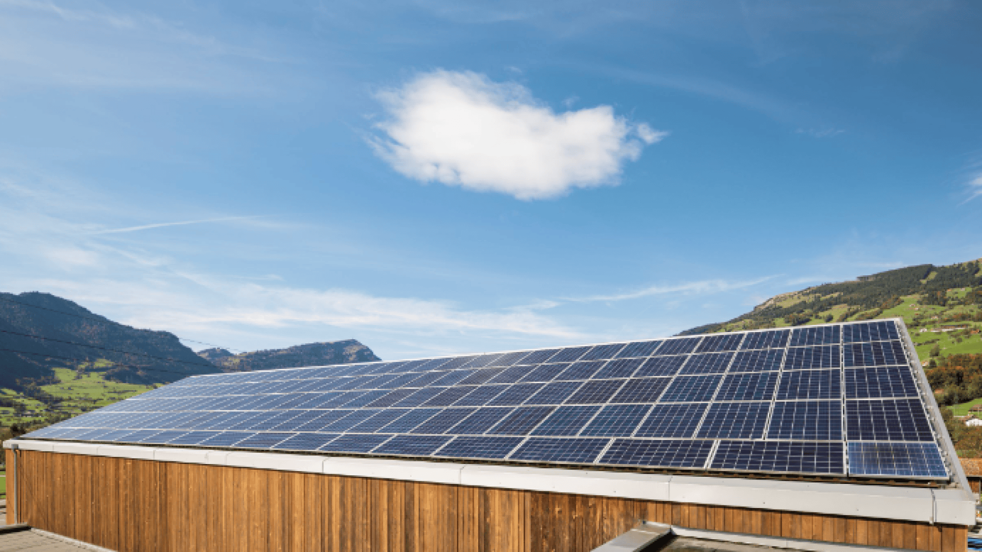 Auf einem Holzdach installierte Solarmodule mit grünen Hügeln und Bergen im Hintergrund unter einem blauen Himmel mit einer einzigen weißen Wolke.