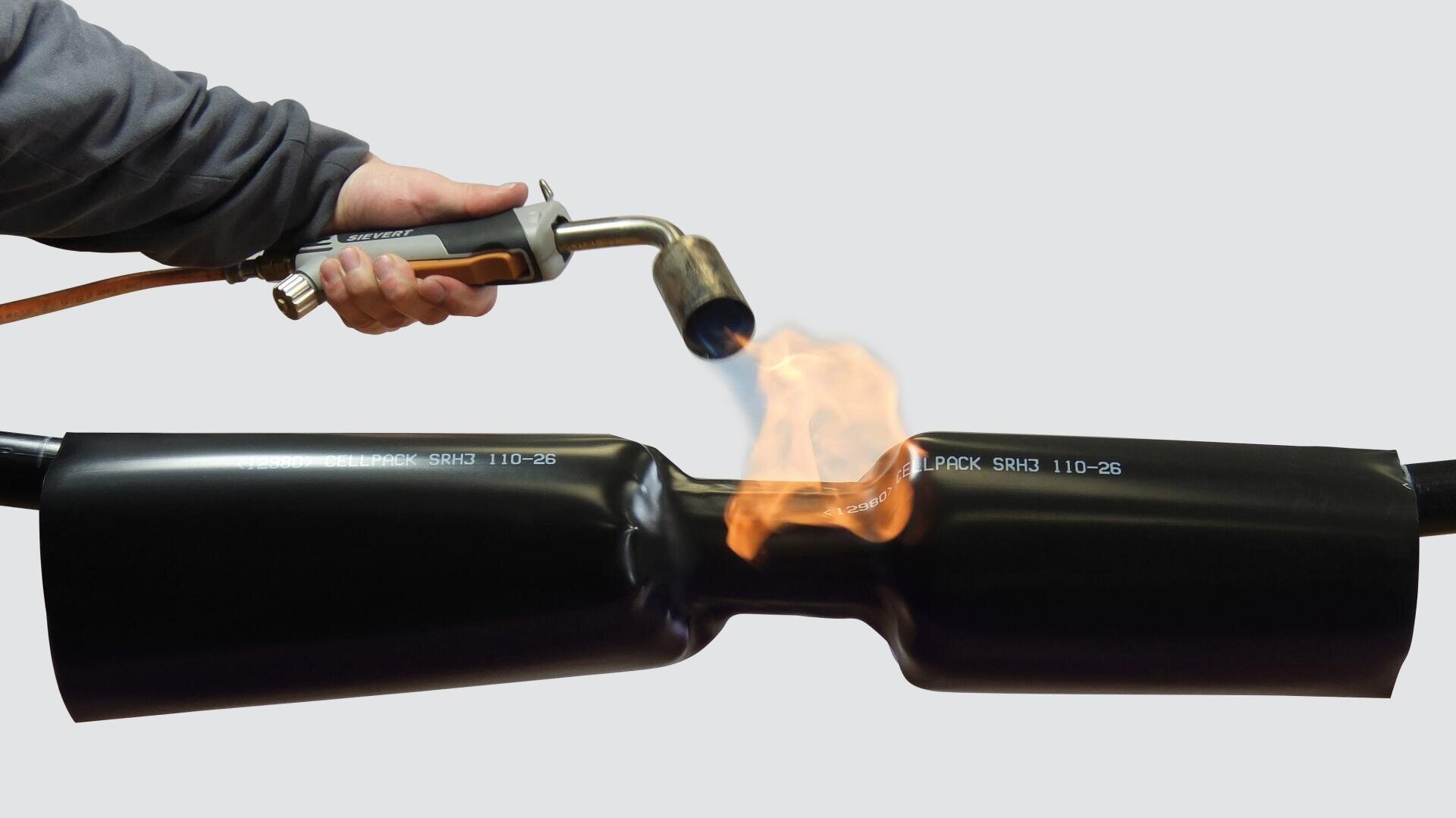 Eine Person erhitzt und schrumpft mit einer Lötlampe einen schwarzen Schrumpfschlauch um ein Rohr, so dass sich das Material unter dem Einfluss der Flammen eng an die Form des Rohrs anpasst.