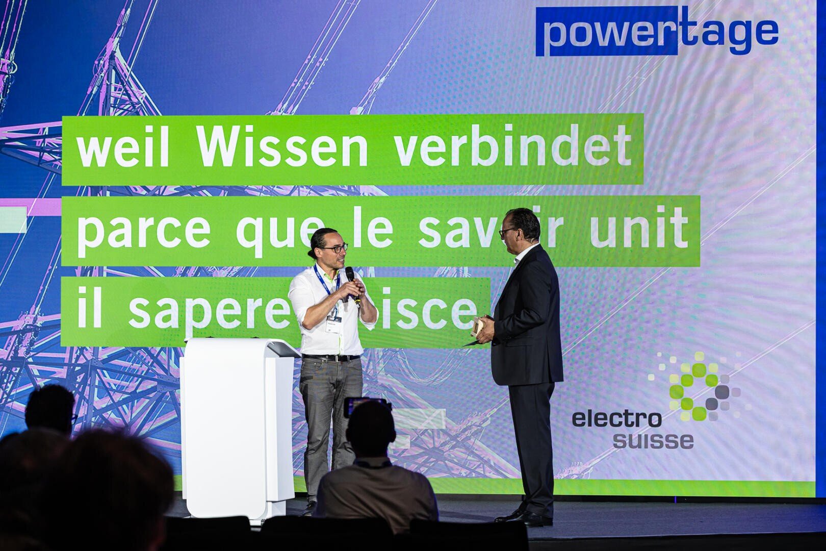 Zwei Männer sprechen auf der Bühne an einer Konferenz. Auf einem Grossbildschirm hinter ihnen sind powertage und der Satz weil Wissen verbindet in Deutsch, Französisch und Italienisch zu sehen, dazu das electrosuisse-Logo.