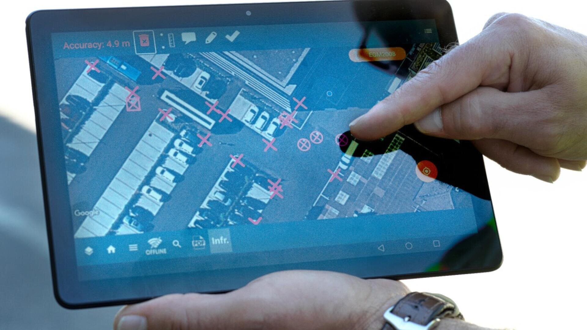 A person uses a tablet displaying an aerial map with marked locations and several pink X marks, possibly indicating key points or vehicles in a parking lot.