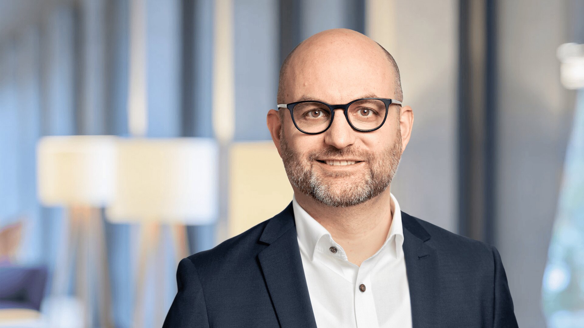 Ein lächelnder Mann mit Brille, gestutztem Bart und einer Glatze trägt einen dunklen Anzug und ein weißes Hemd. Der Hintergrund ist leicht unscharf, moderne Lampen und große Fenster sind zu sehen.