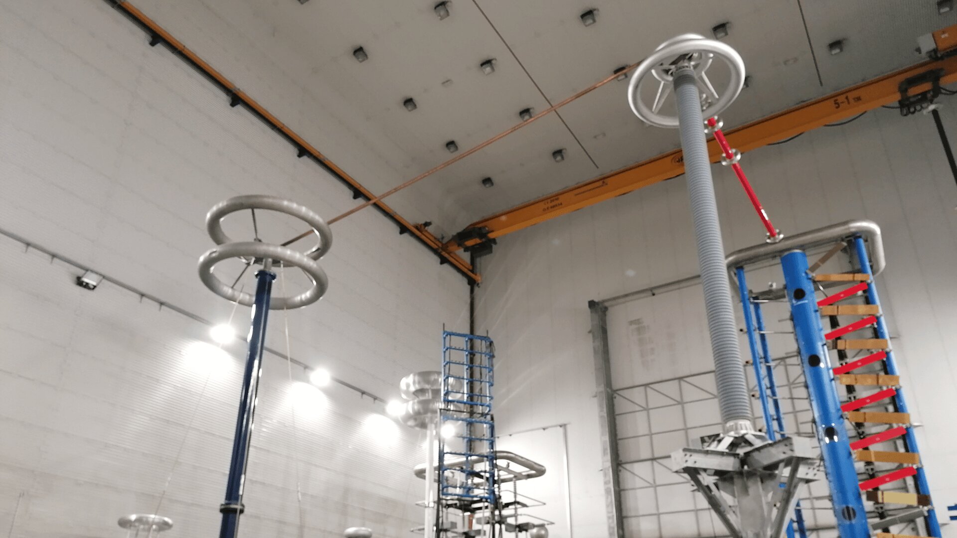 High-voltage test equipment with large metal rings and insulators inside a spacious industrial lab with a crane, lights, and ladders visible on the walls and ceiling.