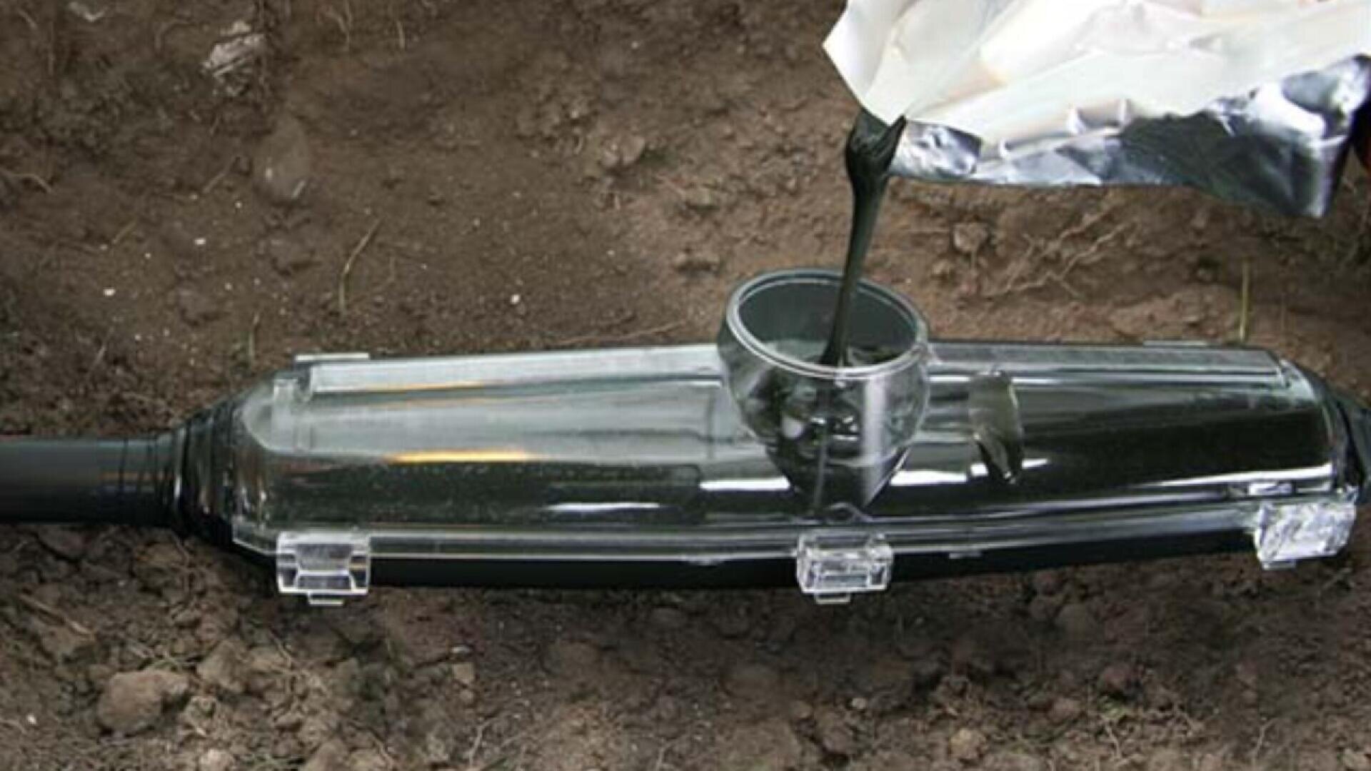A close-up of black liquid being poured from a silver packet into a transparent plastic mold, lying on soil, likely as part of an underground cable joint sealing process.