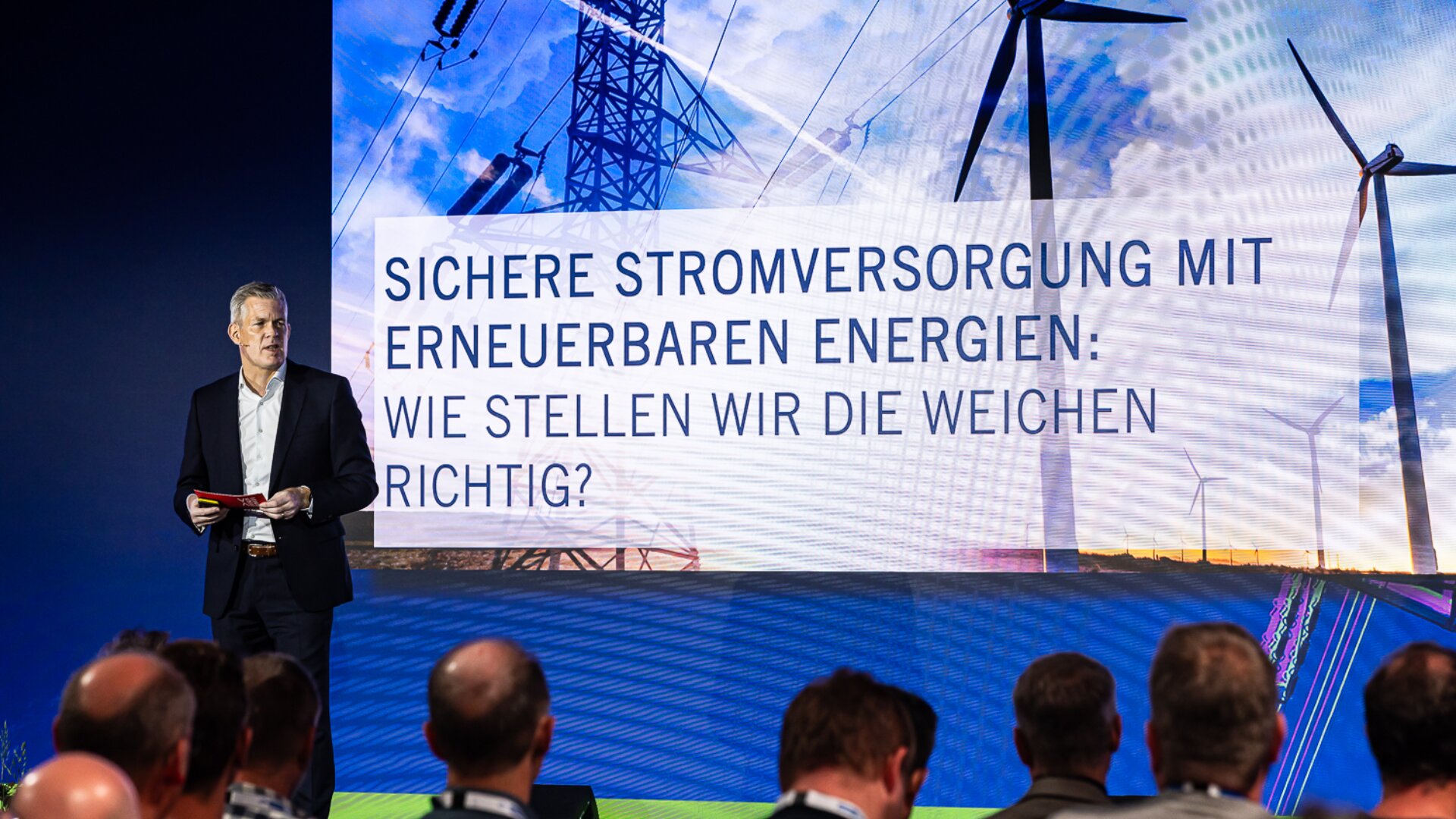 Ein Mann steht auf der Bühne und spricht zu einem Publikum. Hinter ihm wird auf einem großen Bildschirm ein deutscher Text über die sichere Stromversorgung mit erneuerbaren Energien angezeigt, daneben Bilder von Windrädern und Stromleitungen.