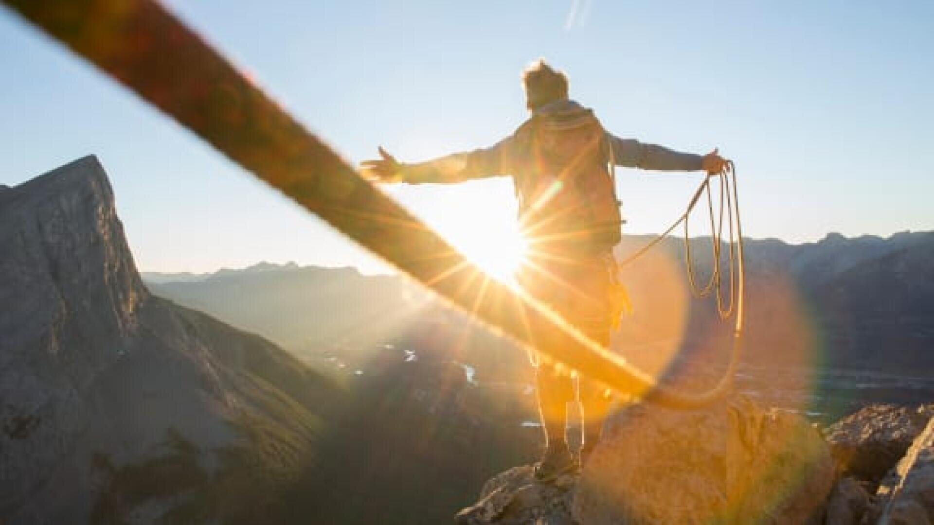 Eine Person steht bei Sonnenaufgang auf einem Berggipfel, die Arme ausgestreckt und mit einem Kletterseil in der Hand, während hinter ihr die Sonne hell scheint und in der Ferne die Berge zu sehen sind.
