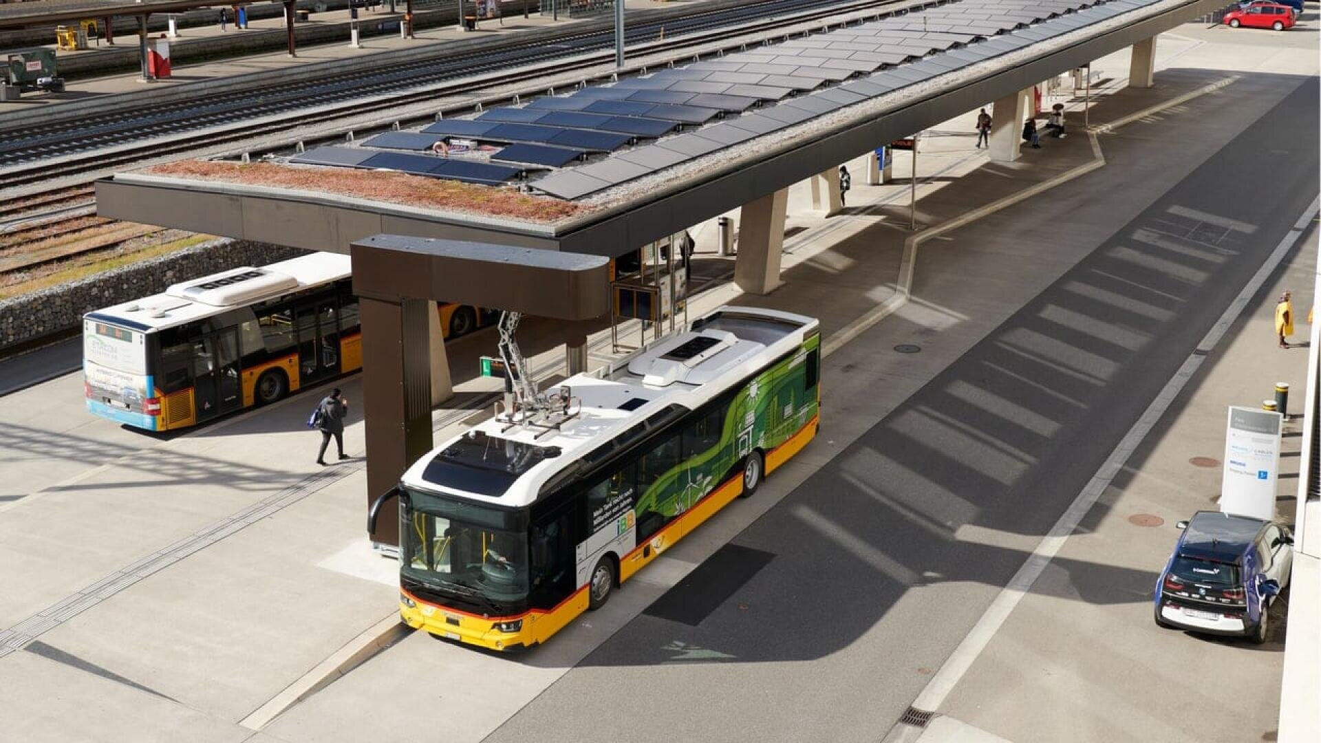 A white and yellow electric bus charges at a bus terminal using an overhead pantograph, with another bus, parked cars, and train tracks visible in the background. A few people walk nearby on the platform.