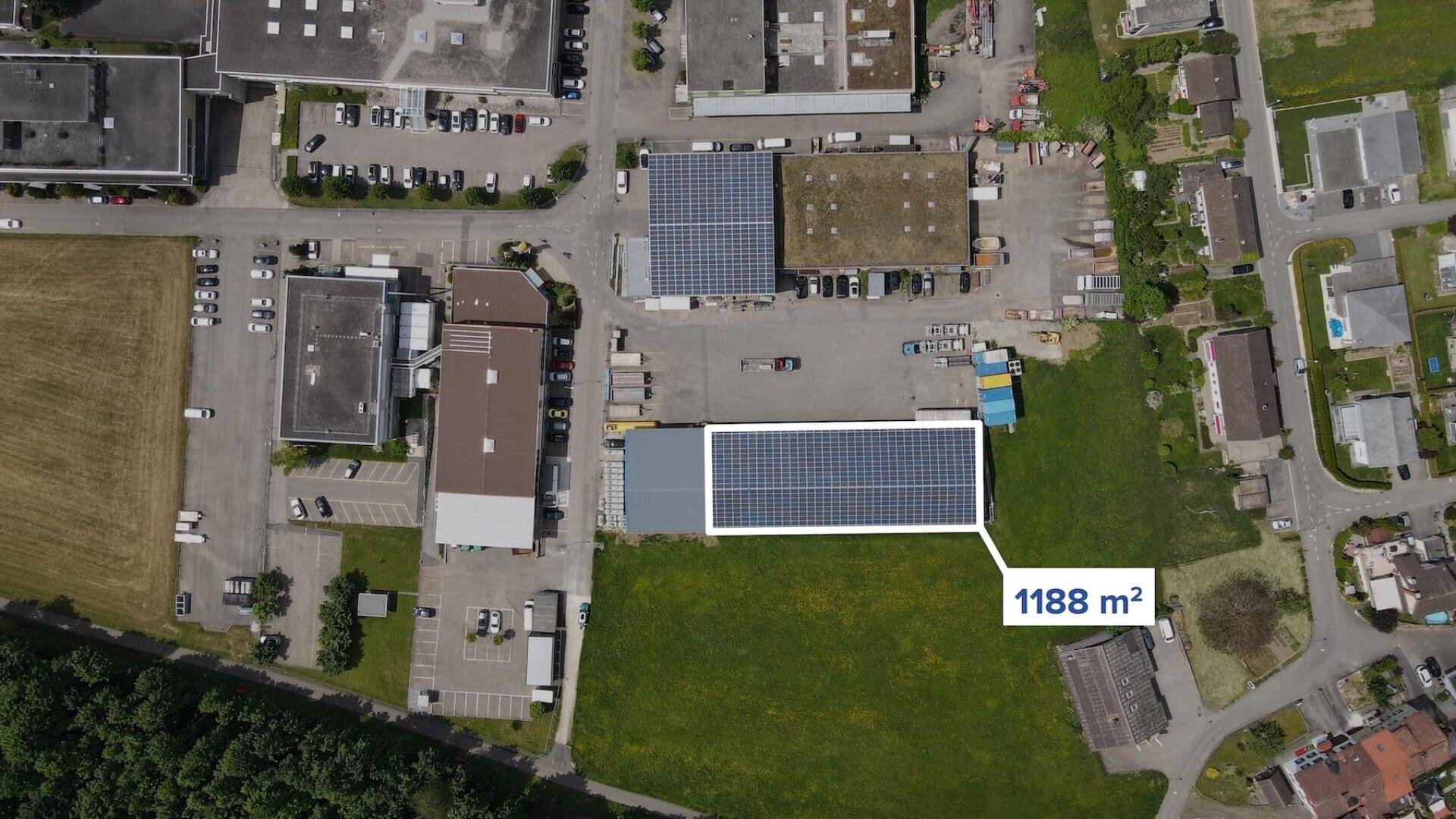 Luftaufnahme eines Industriegebiets mit Gebäuden und Parkplätzen. Es ist ein rechteckiges Gebäude mit Solarzellen auf dem Dach zu sehen, das mit 1188 m² gekennzeichnet ist und an eine grüne Wiese und Wohnhäuser angrenzt.