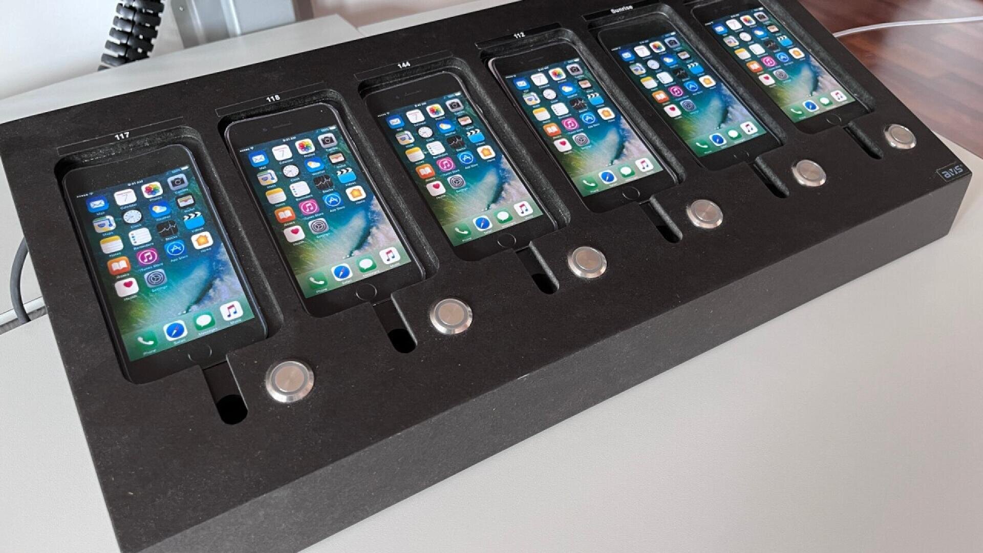 A black charging dock holds six iPhones in a row, each displaying a home screen with app icons. Circular buttons are positioned below each phone slot. The setup is on a white surface.