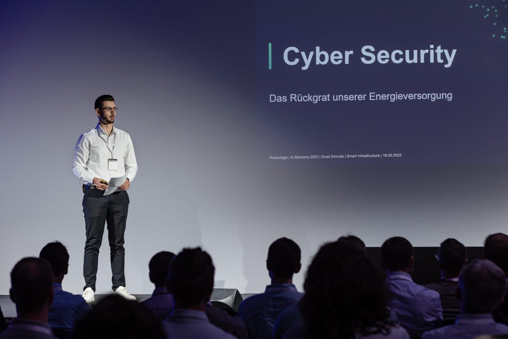 Ein Mann steht auf der Bühne und hält einen Vortrag vor dem Publikum. Hinter ihm ist ein Bildschirm mit dem Titel Cyber Security und einem deutschen Text zum Thema Energieversorgung zu sehen.