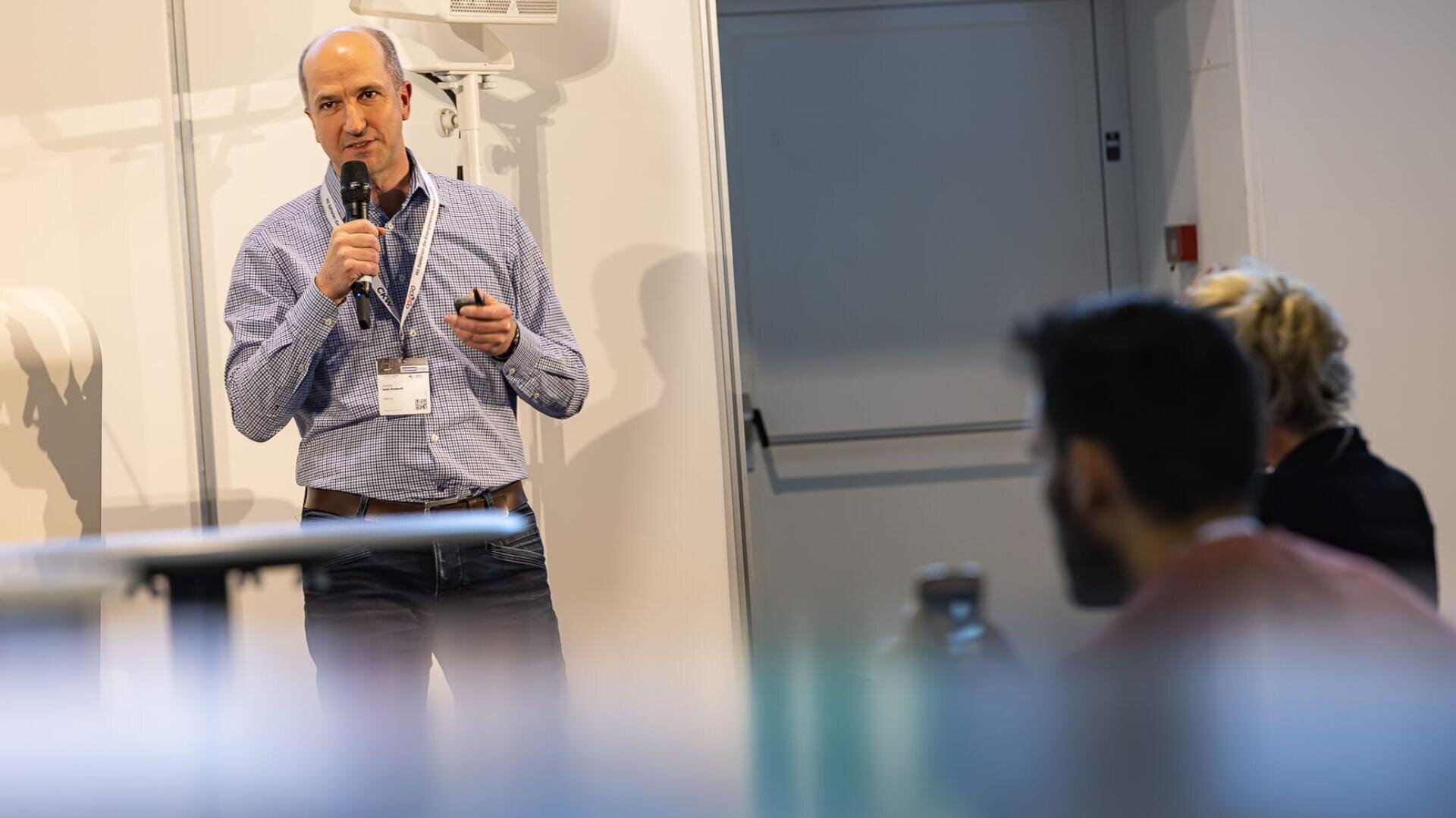 Ein Mann mit Mikrofon und Fernbedienung spricht auf einer Konferenz oder einem Seminar in der Nähe eines Whiteboards. Im Vordergrund sind zwei Zuhörer zu sehen, die aufmerksam zuhören.