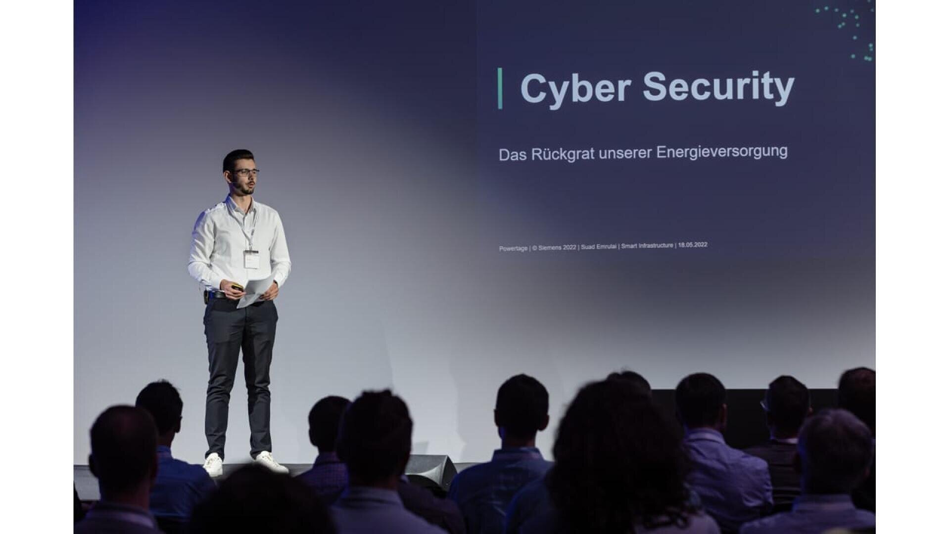 A man stands on stage giving a presentation about cyber security in German to an audience. A large screen behind him displays the title “Cyber Security” and text, with people watching in the foreground.