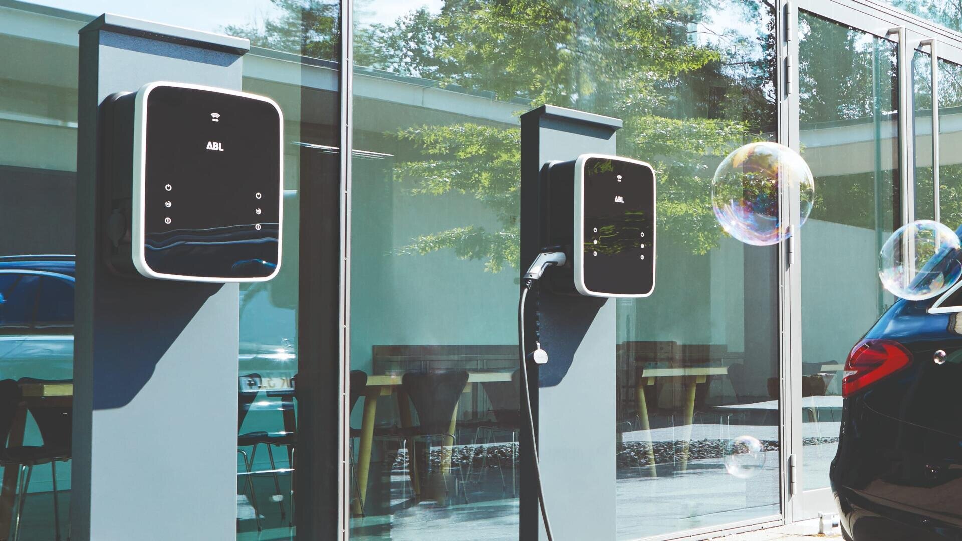 Zwei Ladestationen für Elektrofahrzeuge sind an Pfosten außerhalb eines Glasgebäudes angebracht. Eine Station ist in Betrieb und lädt ein schwarzes Auto auf. Auf den Glasfenstern sind Spiegelungen von Bäumen und großen Luftblasen zu sehen.