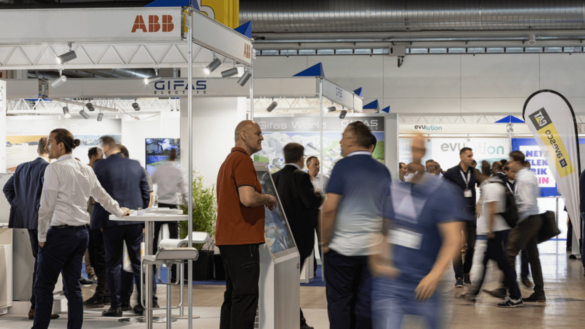 Auf einer belebten Indoor-Messe oder -Ausstellung laufen und reden die Menschen, umgeben von Ständen mit Firmenbannern und Displays. Einige Teilnehmer tragen Namensschilder und Geschäftskleidung und beteiligen sich an Netzwerken und Diskussionen.