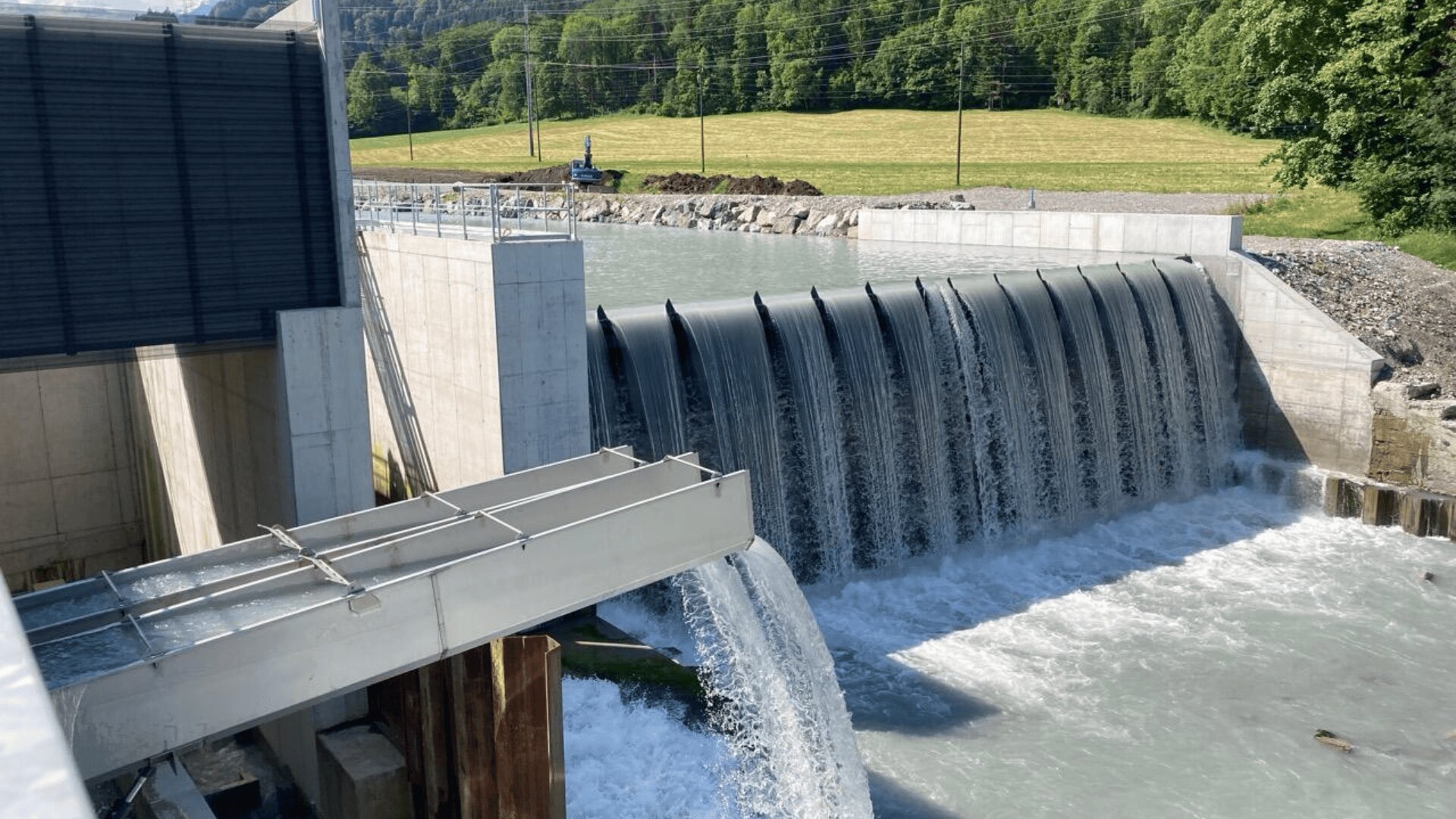 Ein moderner Staudamm mit Wasser, das über geschwungene Tore und eine Überlaufrinne fließt, umgeben von Grünflächen und einer Wiese im Hintergrund.