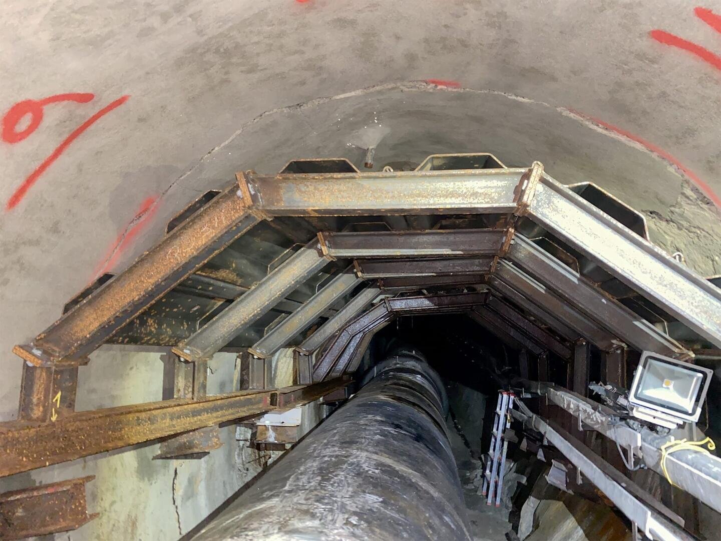 Blick ins Innere eines mit Metallträgern verstärkten Tunnels oder Schachts mit einem großen zentralen Rohr, elektrischen Kabeln, einer angebrachten Lampe und roten Markierungen an der Betonwand. Der Tunnel reicht bis in die Dunkelheit.