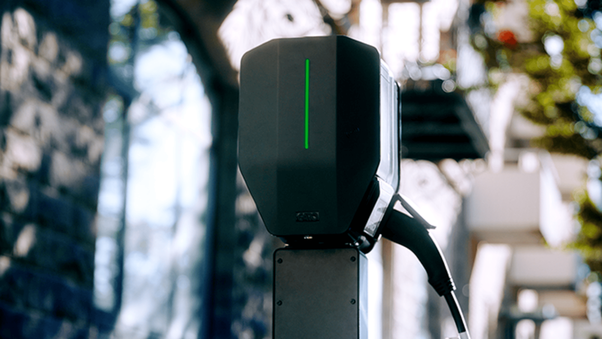Eine Nahaufnahme einer schwarzen Ladestation für Elektrofahrzeuge mit grünem Licht, die im Freien an einem Pfosten montiert ist, vor einem unscharfen Hintergrund aus einer Steinmauer und Bäumen.