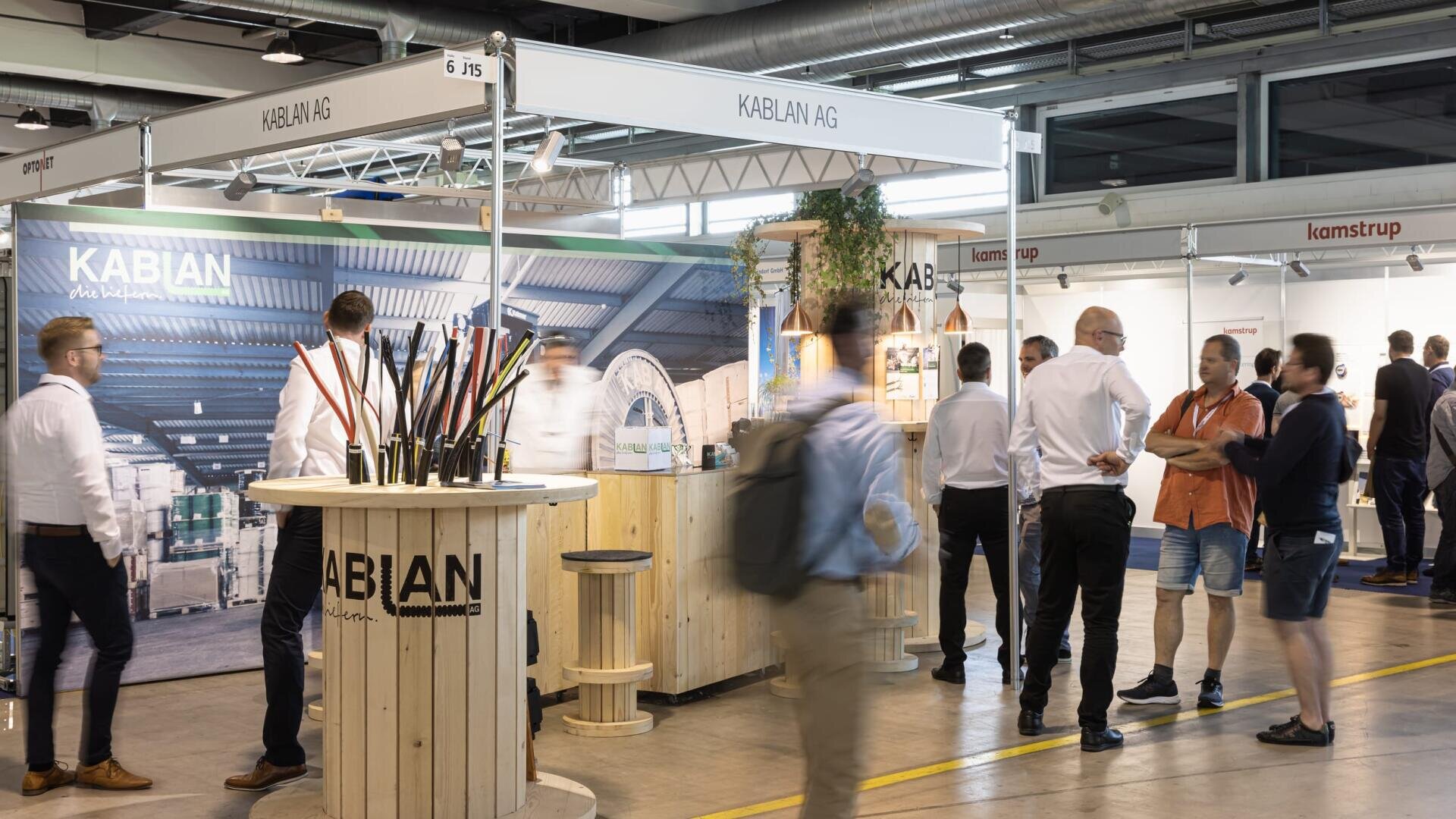 A busy trade show with people talking and walking near the KABLAN AG booth, which features wooden displays and a table with cable samples. Other exhibition stands and banners are visible in the background.