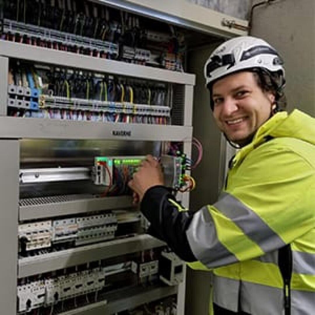 Ein lächelnder Elektriker, der einen Helm und eine Warnweste trägt, arbeitet an einer offenen elektrischen Schalttafel, die mit Drähten und Leistungsschaltern gefüllt ist.
