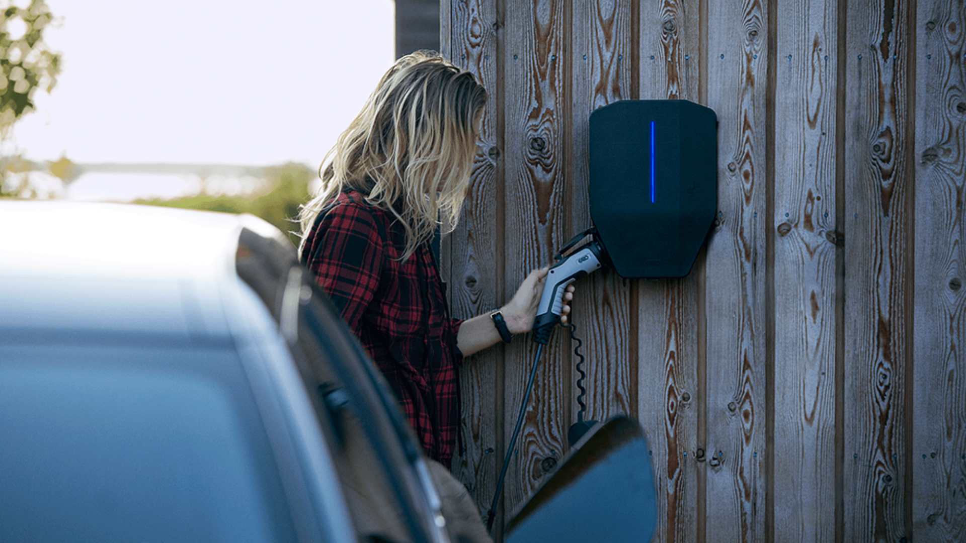Eine Person mit langen Haaren und einem rotkarierten Hemd schließt ein Ladekabel an eine Ladestation für Elektrofahrzeuge an, die an einer Holzwand angebracht ist, wobei im Vordergrund ein Auto teilweise zu sehen ist.