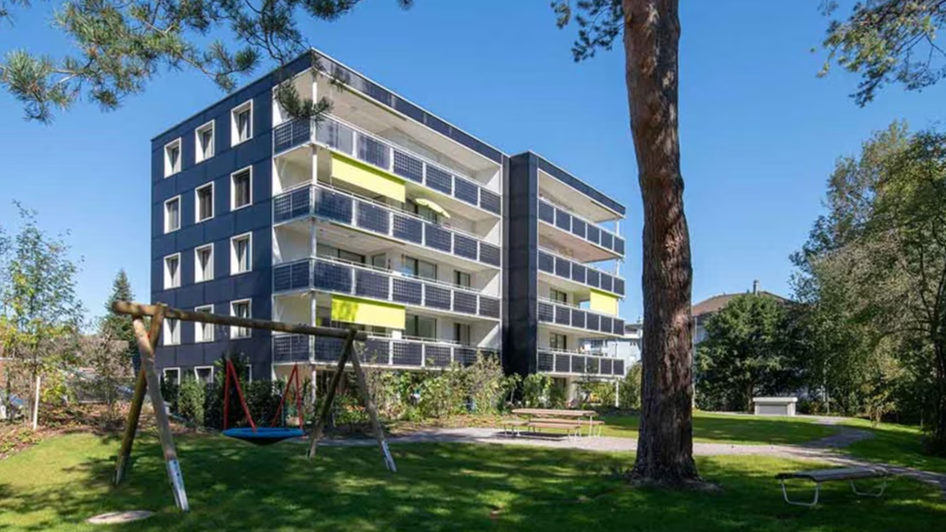 Ein modernes vierstöckiges Wohngebäude mit Balkonen, umgeben von Bäumen und grünem Gras. Im Vordergrund gibt es eine Schaukel, Picknicktische und einen Weg, alles unter einem klaren blauen Himmel.