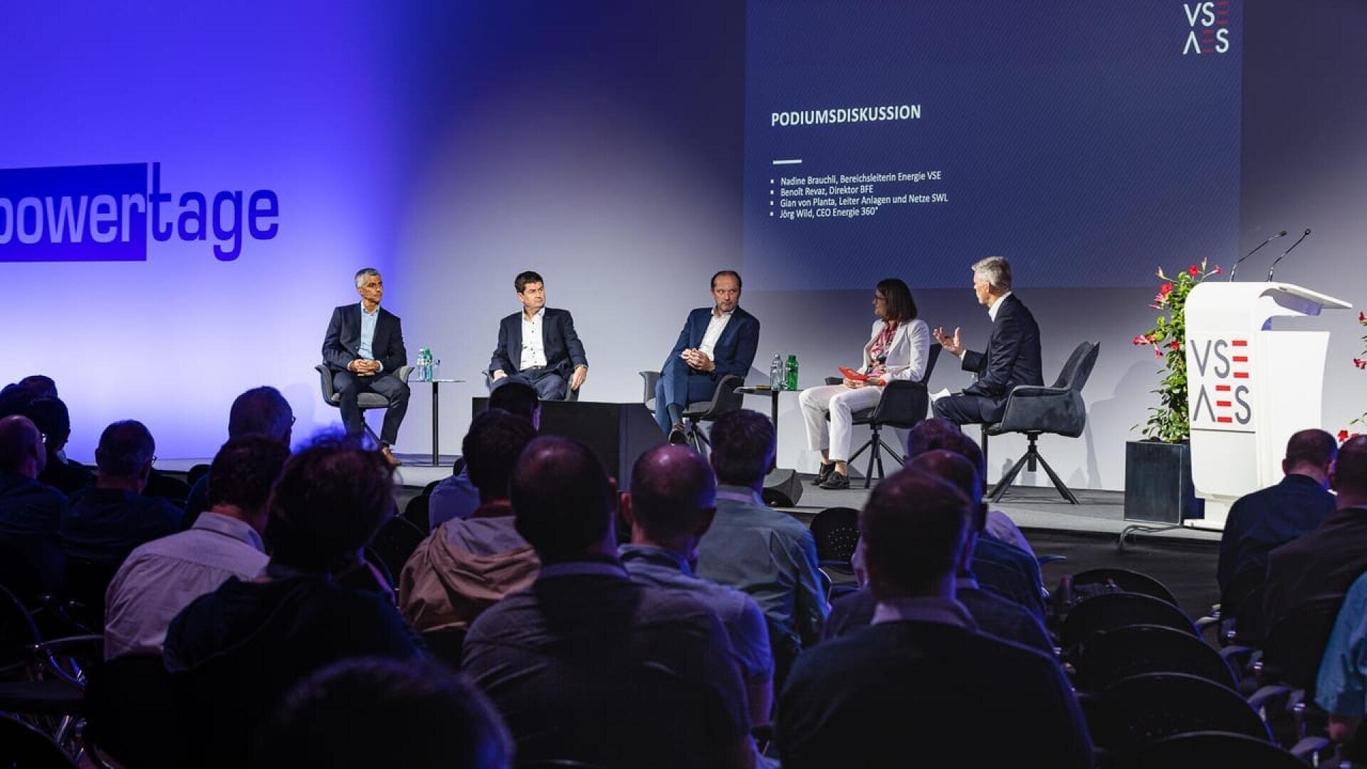Five panelists sit on stage having a discussion in front of an audience at a conference. A screen behind them displays text with discussion details. The atmosphere is professional, with branded decorations and lighting.
