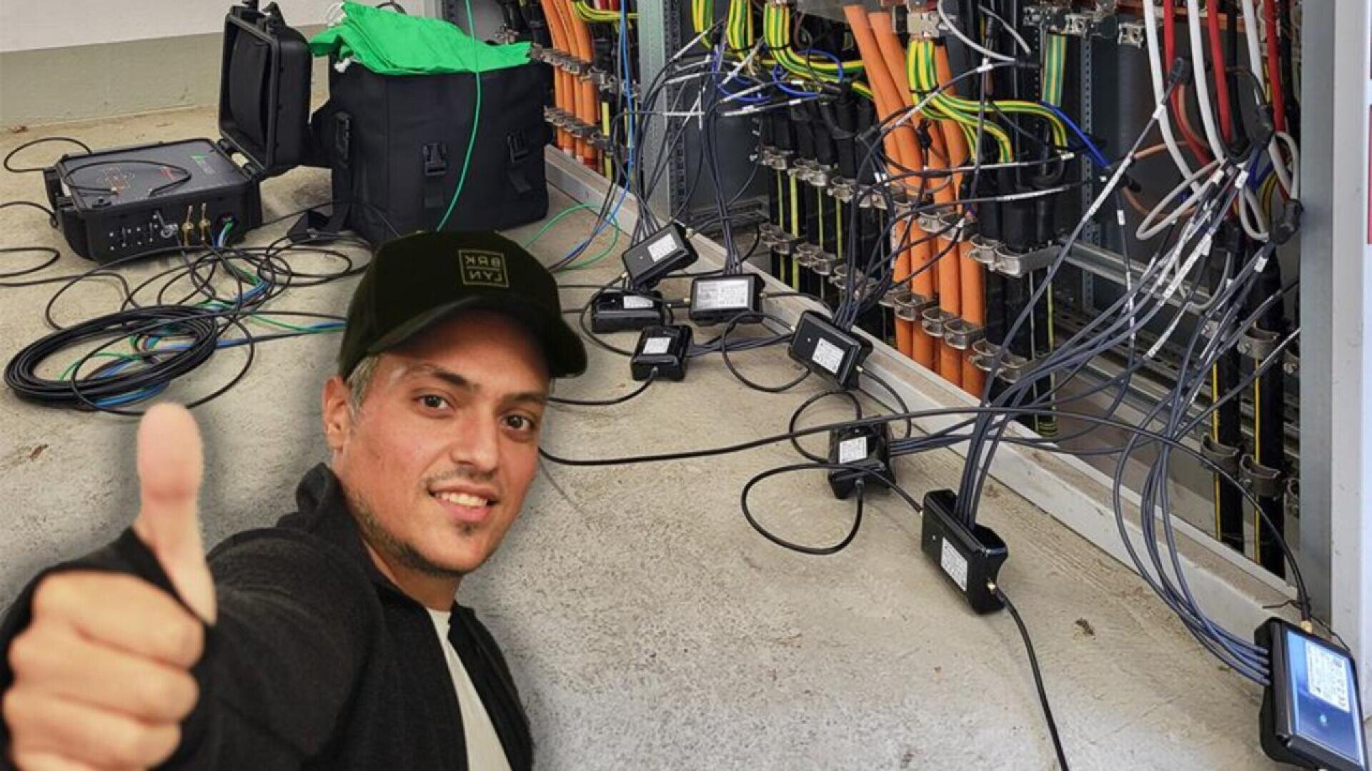 A man in a black cap gives a thumbs up in front of an open electrical panel with numerous exposed wires, cables, and electronic equipment on the floor.