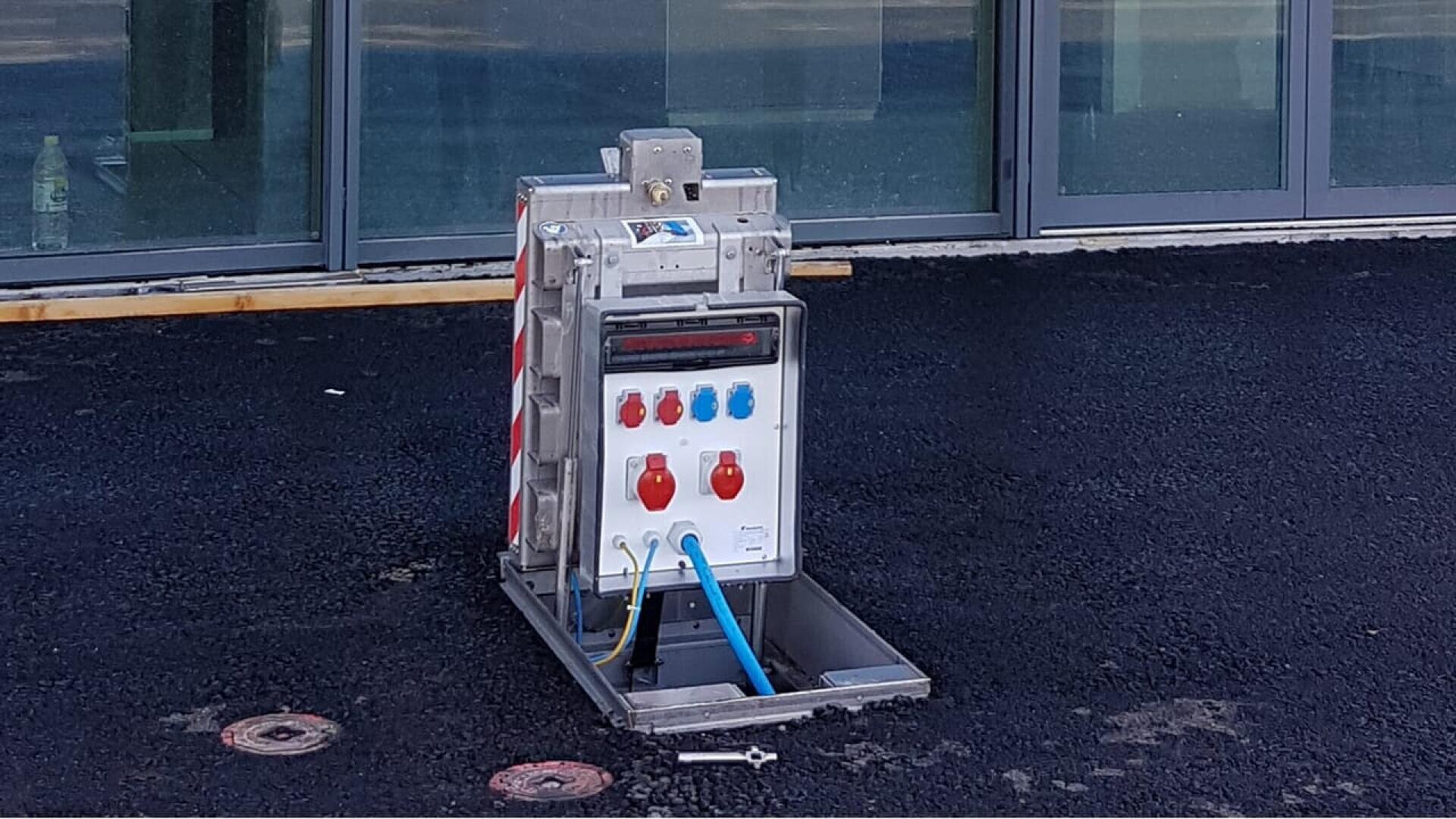 A metal outdoor electrical box with multiple colored sockets and cables is installed in the asphalt ground in front of glass windows. The cover is open, and tools lie nearby.