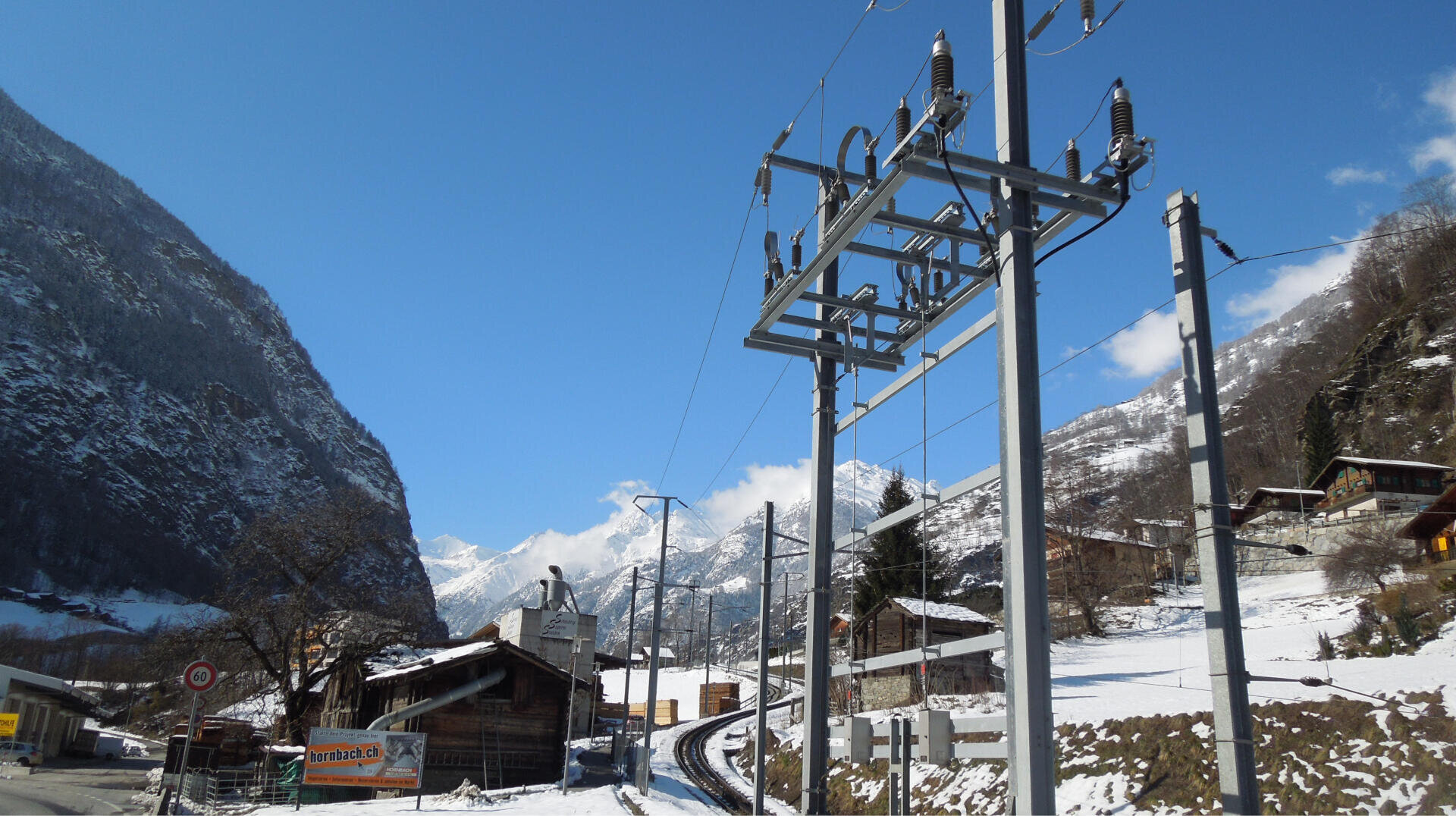 Ein verschneites Dorf in einem Gebirgstal mit Holzhäusern, Stromleitungen, Strommasten und einem strahlend blauen Himmel. Auf beiden Seiten erheben sich felsige, bewaldete Berge, und eine verschneite Straße schlängelt sich durch die Szenerie.