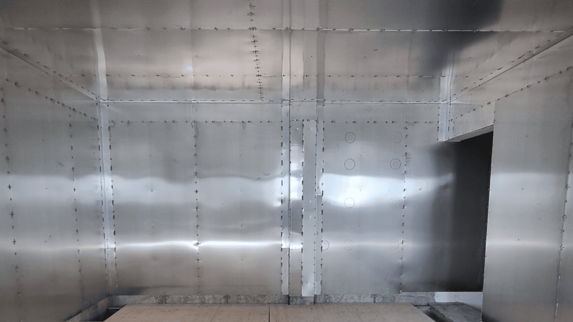 Ein großer, leerer Raum mit glatten, spiegelnden Metallwänden mit sichtbaren Nieten und Nähten, einem Betonboden und einer dunklen Tür auf der rechten Seite.