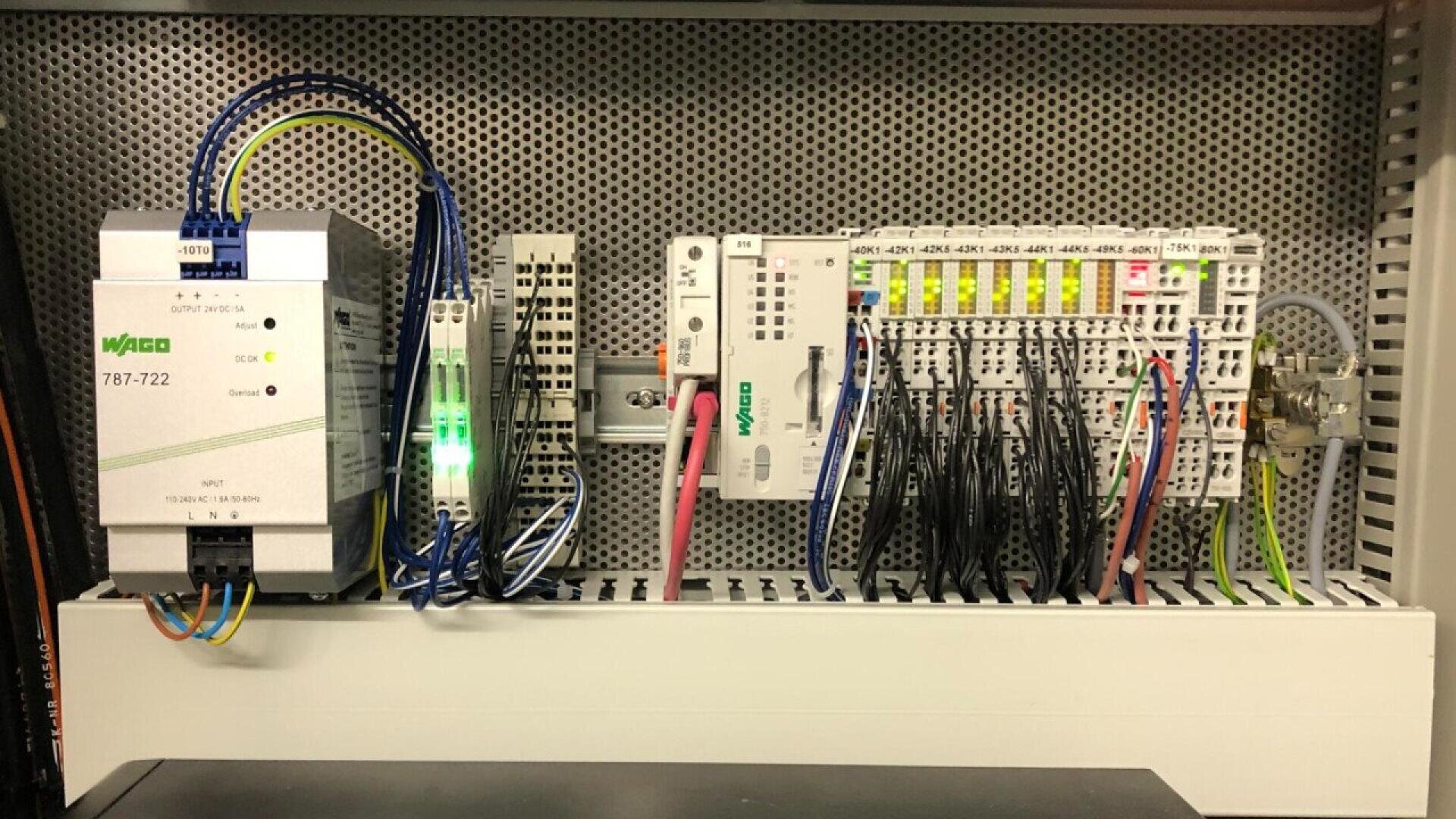 A control panel featuring various electronic components and wiring, including a WAGO 787-722 power supply, terminal blocks, relays, and connected cables, all mounted on a perforated metal backplate.
