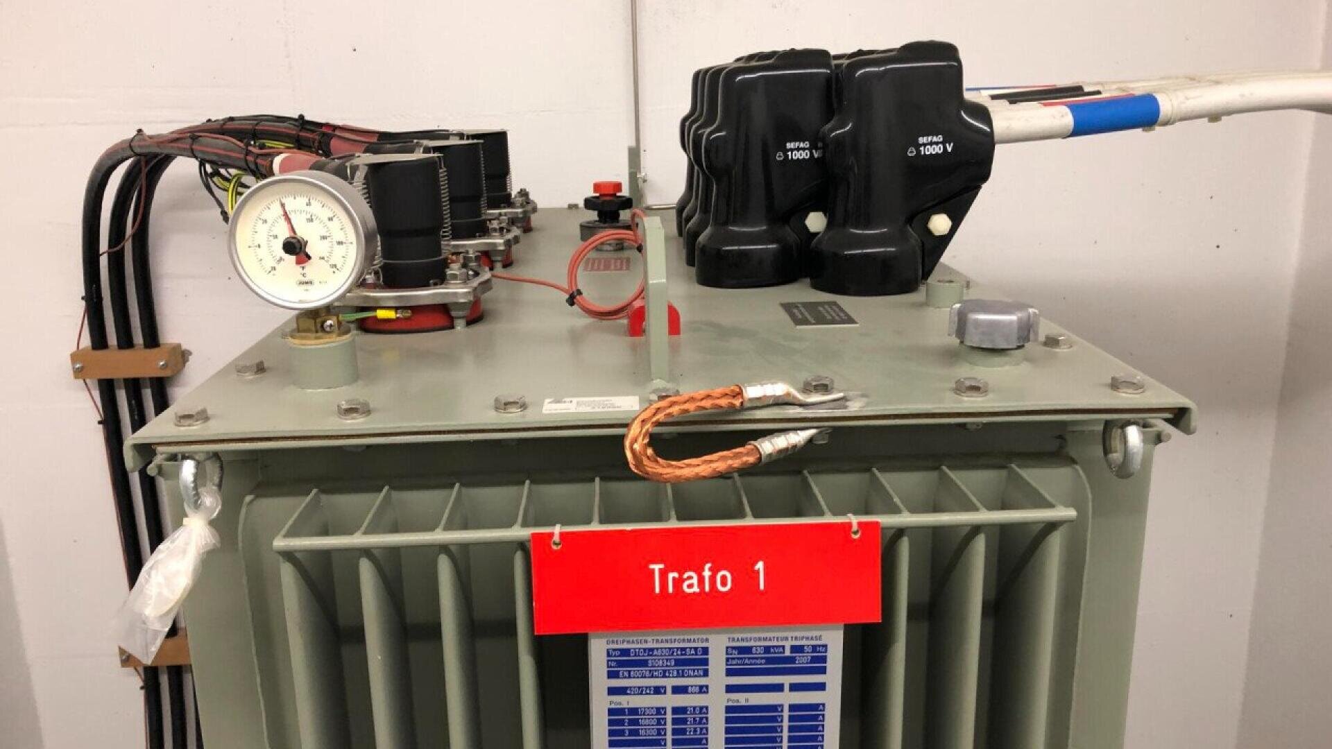 A close-up view of an electrical transformer labeled Trafo 1 with cables, gauges, and connectors visible on top; a red sign hangs on the front of the gray metallic unit.