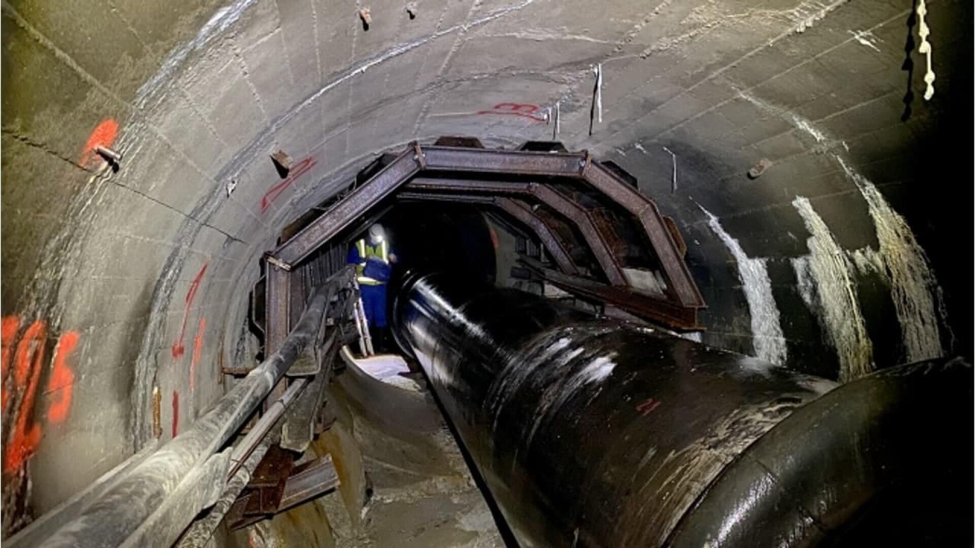 A worker in a reflective vest stands inside a large, dimly lit concrete tunnel with metal reinforcements and a large black pipe running through the center. Red markings are visible on the tunnel walls.