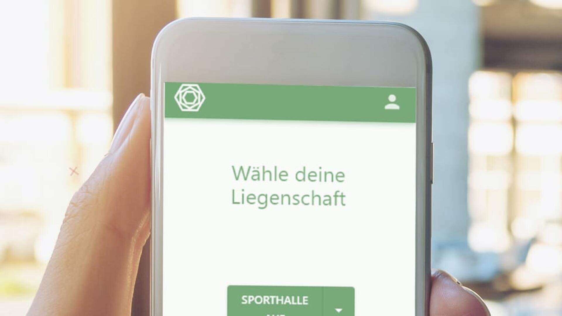 Eine Hand hält ein Smartphone, das einen grün-weißen App-Bildschirm mit dem deutschen Text Wähle deine Liegenschaft und einer Taste mit der Aufschrift Sporthalle anzeigt. Der Hintergrund ist sanft verschwommen.